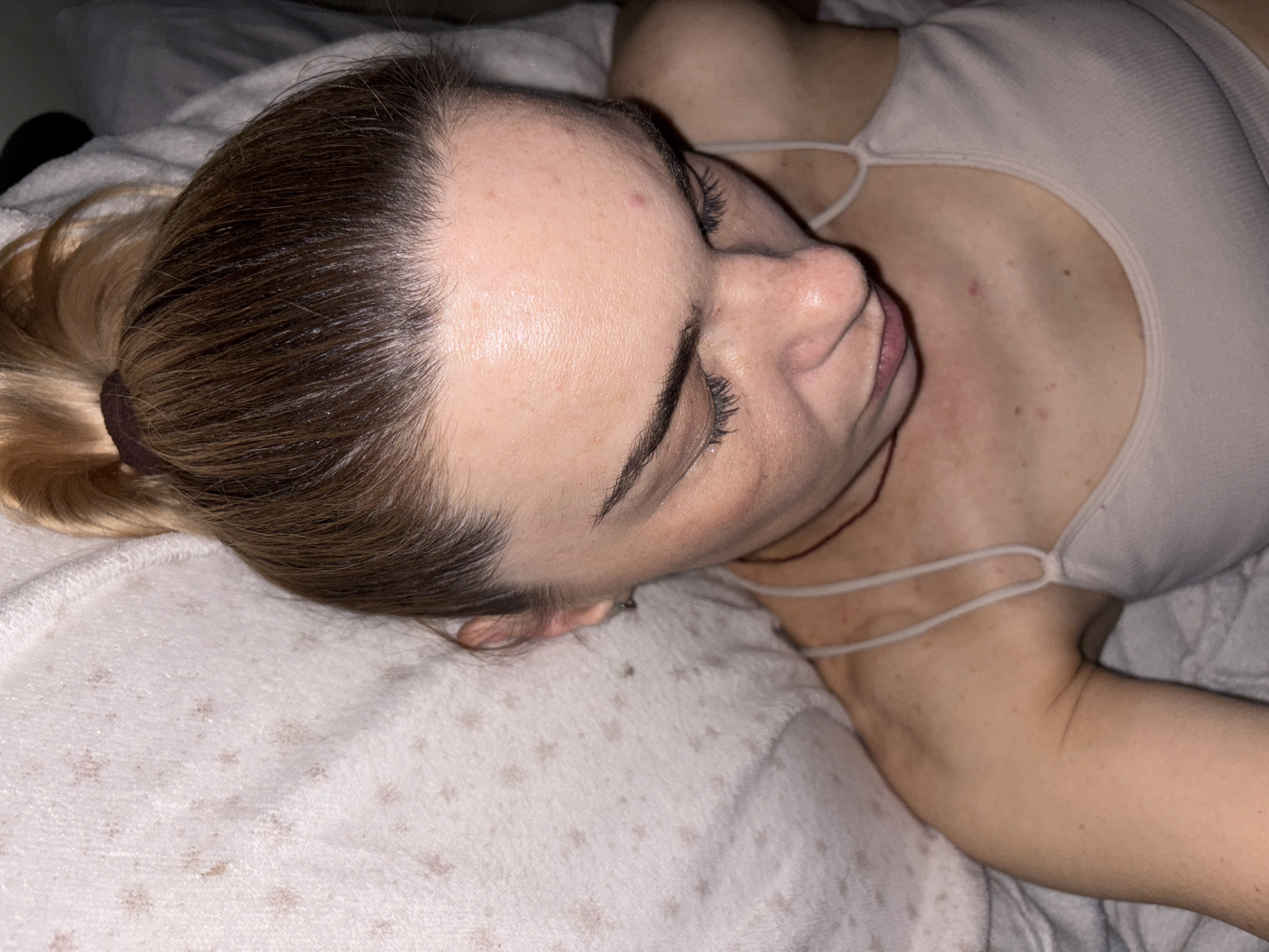 A close-up overhead shot shows a young woman lying down, looking down. Her hair is pulled back in a high ponytail. She is wearing a light-colored tank top. The background is a light-colored blanket with a subtle star pattern. The environment is indoors. The subject is alone in the frame. There are no visible landmarks or contextual clues to suggest a specific location in a city or country.