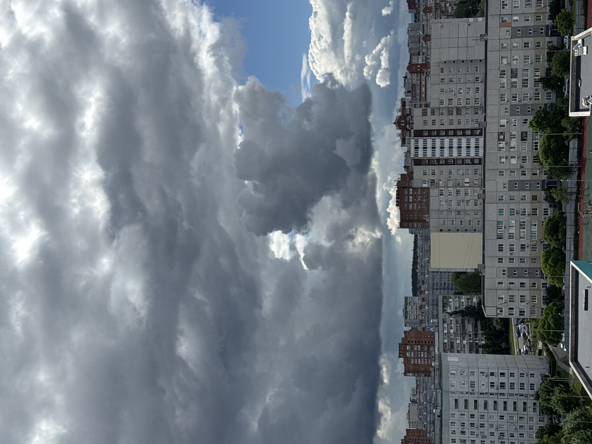 The image displays a panoramic view of an urban landscape under a dramatic sky, presented as if the horizon is on the right side due to vertical camera orientation. When viewed correctly, the upper two-thirds of the image are dominated by a tumultuous sky, filled with a complex interplay of dark, heavy grey clouds, bright white, sunlit cloud formations, and occasional glimpses of clear blue sky. This dynamic cloud cover creates a striking contrast of light and shadow, suggesting a changeable weather pattern, possibly an approaching or receding storm.
The lower third of the image features a dense collection of multi-story apartment buildings, typical of Eastern European or former Soviet-bloc architecture. These buildings vary in color, predominantly grey concrete and reddish-brown brick, with numerous windows. Many of the buildings have air conditioning units protruding from their windows. Green trees and other foliage are visible between the structures, providing some natural contrast to the built environment. On the far right, in the foreground, a corner of what appears to be a sports court (possibly tennis or basketball) can be seen. Streets with a few visible cars weave through the urban fabric at ground level. No specific activities or individuals are discernible, and no legible text is visible on any signs or buildings. The overall impression is that of a bustling urban area experiencing a visually remarkable day.