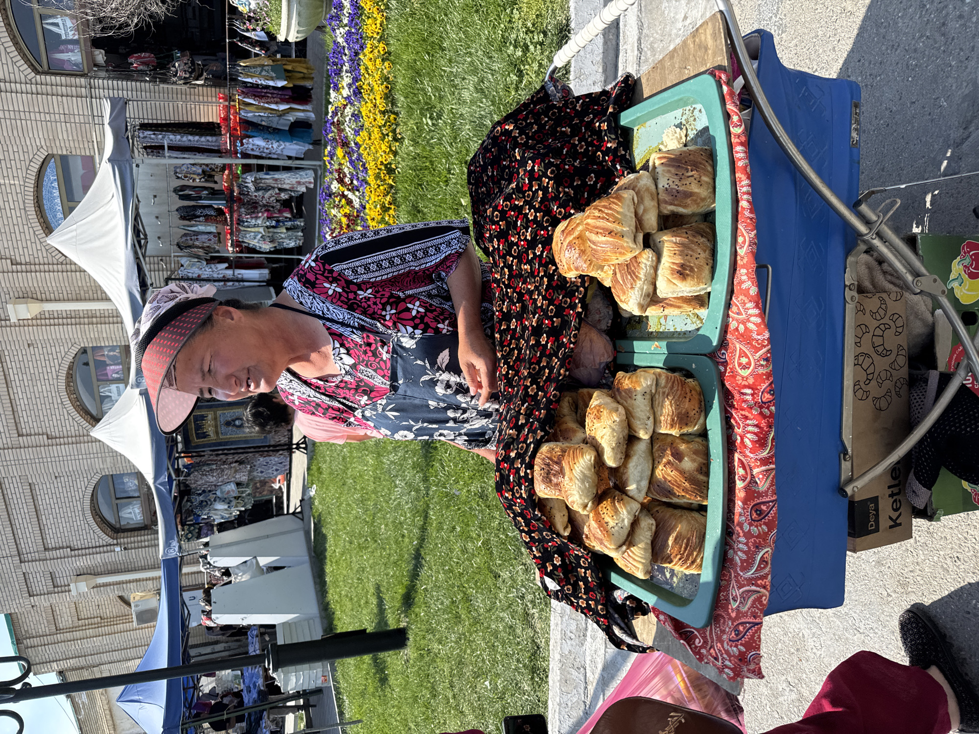 A woman vendor sells baked goods at a food stand in Samarkand City, Uzbekistan, on a sunny day. She stands behind the stand, which is on the sidewalk next to a patch of green grass. There are two green trays, filled with golden-brown pastries, on top of the stand. A black and red floral-patterned scarf partially covers the trays. The vendor is wearing a pink and black patterned top with a black floral apron and a pink visor. In the background are market stalls, clothing racks, a brick building with arched windows, and a flower bed.