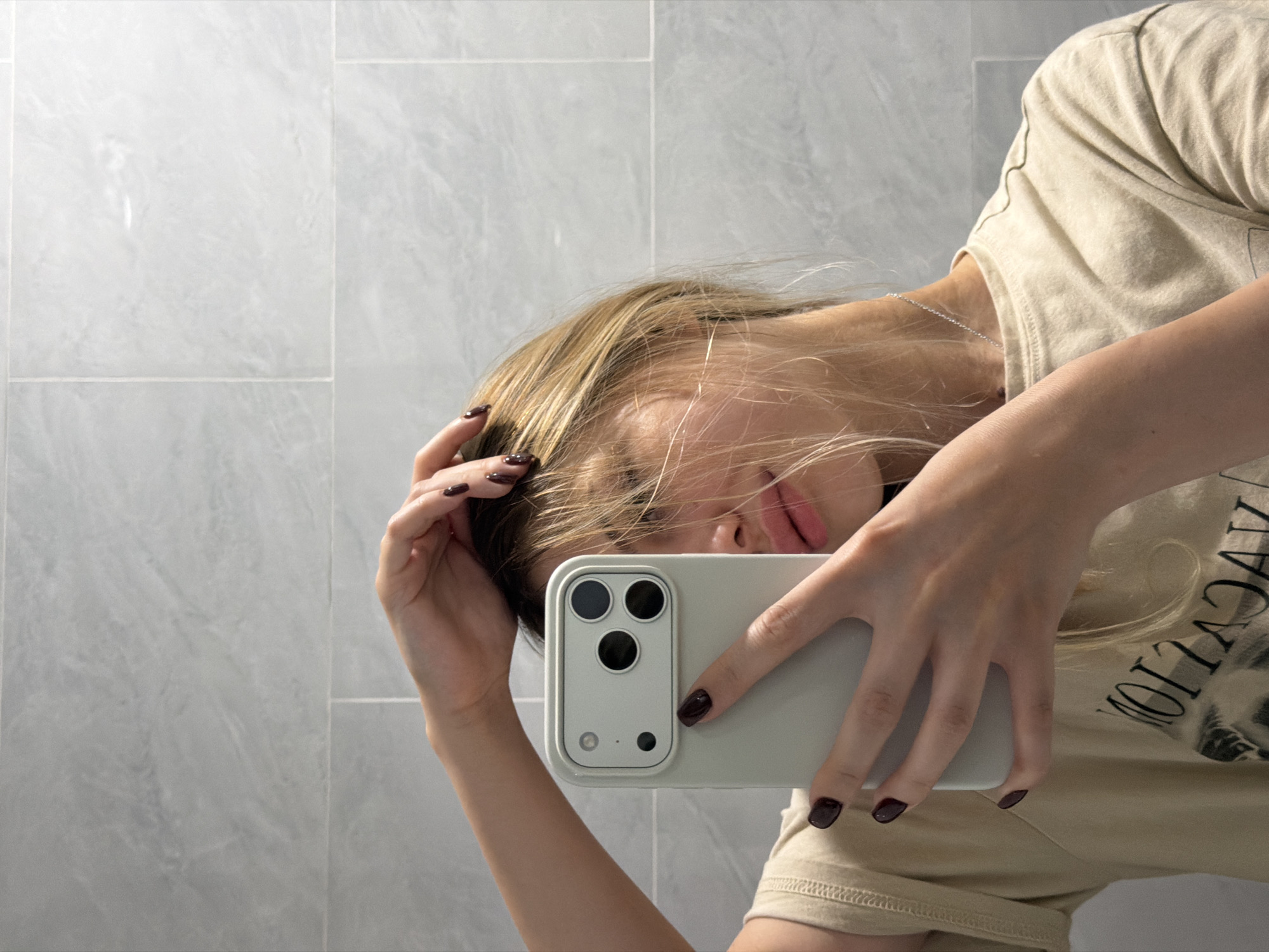 A young woman with blonde hair is taking a selfie in a bathroom with grey tiled walls. She is wearing a light beige t-shirt with a graphic print. Her left hand is holding a light grey smartphone, and her right hand is holding her hair. Her nails are painted a dark burgundy color. The lighting is bright, creating a clean and modern aesthetic. The background shows the grey tiled walls of the bathroom, reflecting the light and creating a minimalist environment. There is no indication of the location being in Da Nang, Vietnam, or any other specific city.