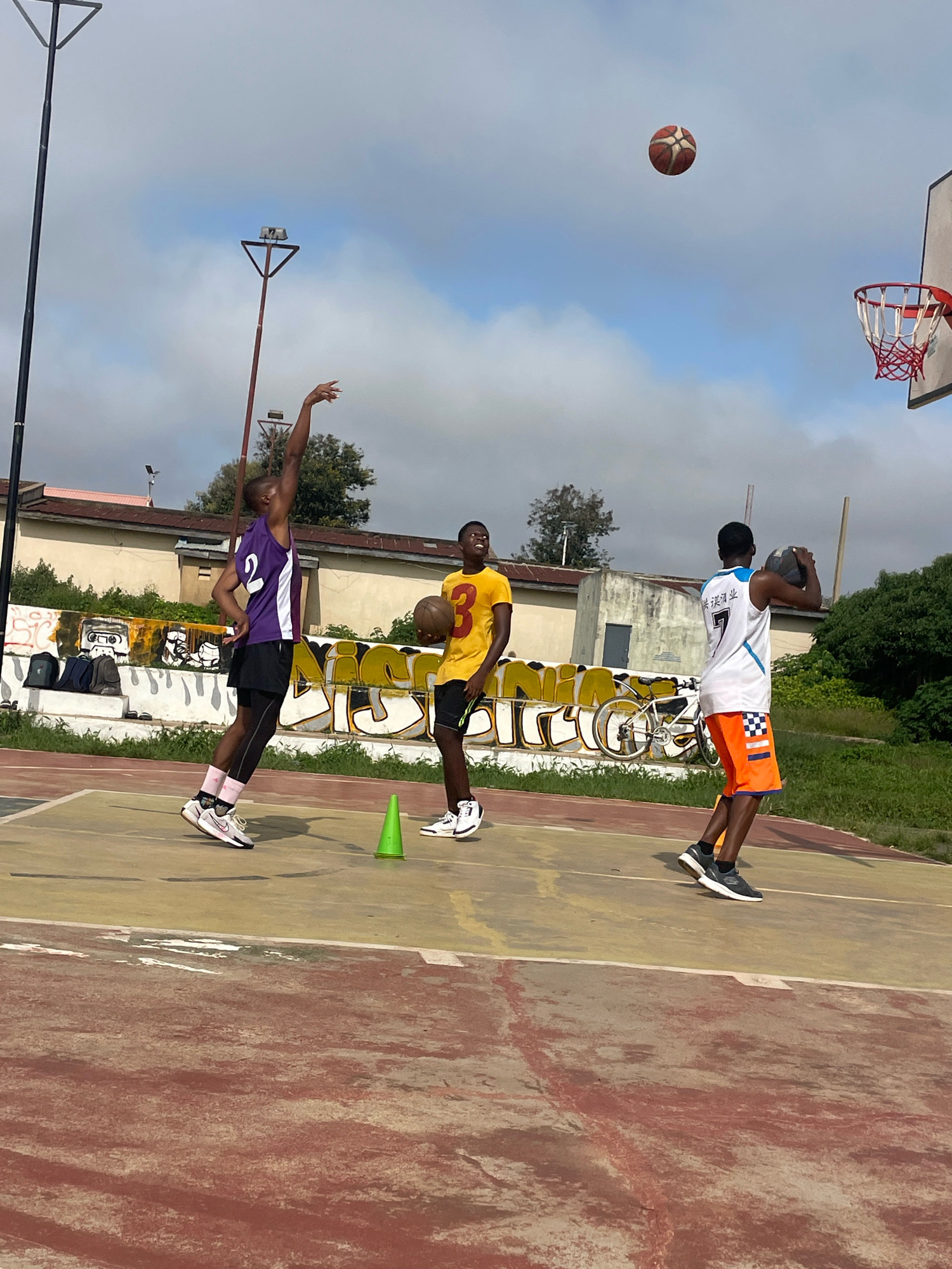 In Gura Top, Nigeria, three young men are playing basketball on an outdoor court. The court surface is a worn, reddish-brown with yellow markings. In the background, there's a low building with graffiti on its lower section, depicting various images and lettering. Two streetlights with triangular tops stand tall. The sky is partly cloudy with a light blue hue.
The person on the left, wearing a purple and white basketball jersey with the number '2' and black shorts, is in mid-air, jumping to shoot a basketball. They are wearing white and pink sneakers. In the middle, a young man in a yellow jersey with the number '3' and black shorts holds a basketball and is looking towards the shooter. He is wearing white sneakers. The person on the right, dressed in a white and blue jersey with the number '7' and bright orange shorts with a checkered pattern, is also holding a basketball and appears to be observing the play. They are wearing dark sneakers. A basketball is visible in mid-air, above and to the right of the shooter, suggesting a shot has just been taken. A green traffic cone is placed on the court near the middle player. Several backpacks are visible on a ledge in the background, next to the graffiti. A white bicycle is leaning against the wall in the middle ground.