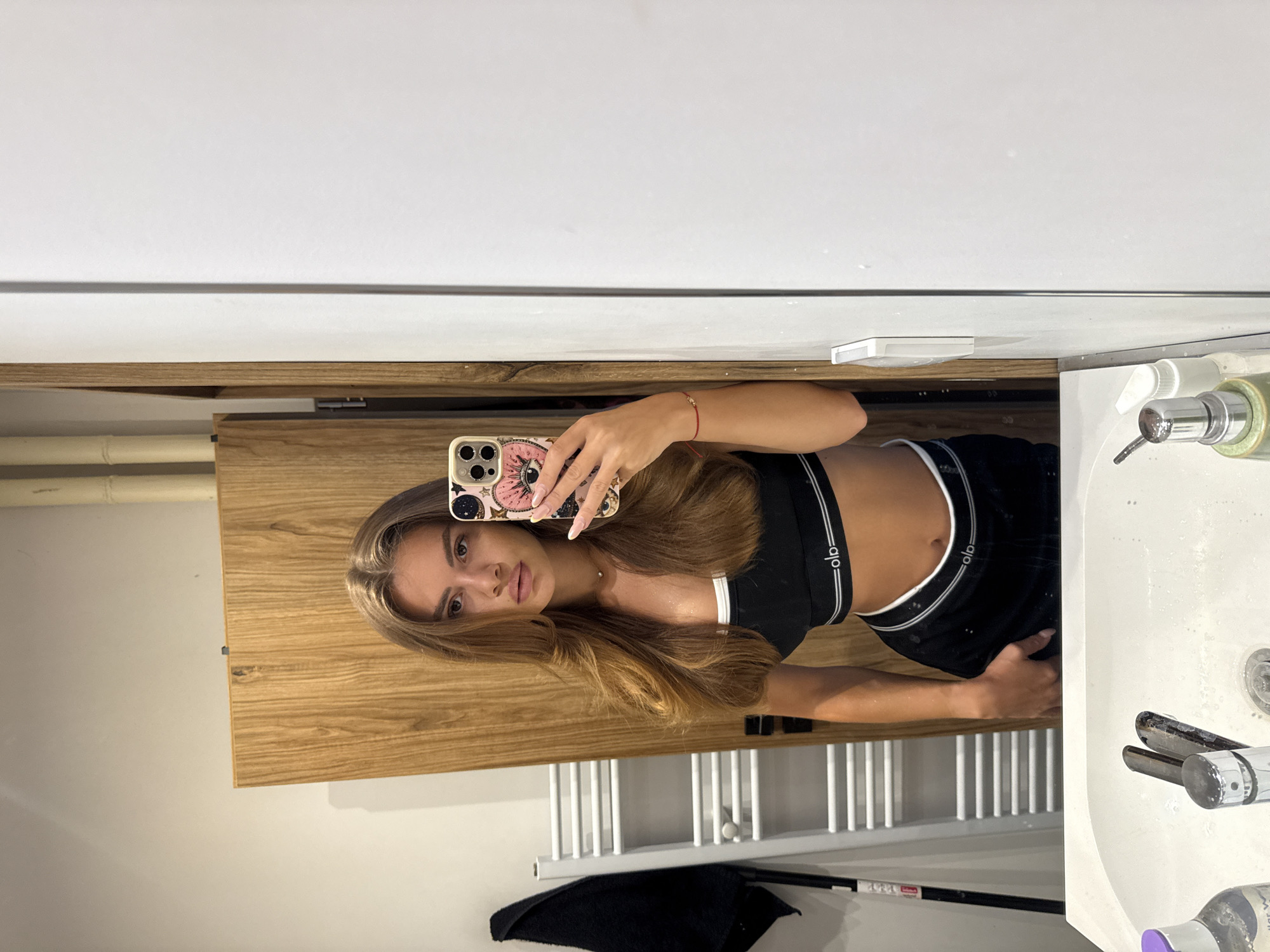 A young woman with long, brown hair takes a mirror selfie in a bathroom. She is wearing a black sports bra and black shorts. The background includes a wooden cabinet, a towel rack with a black towel, and a white sink. The image does not contain any information indicating the location is in Plovdiv, Bulgaria.