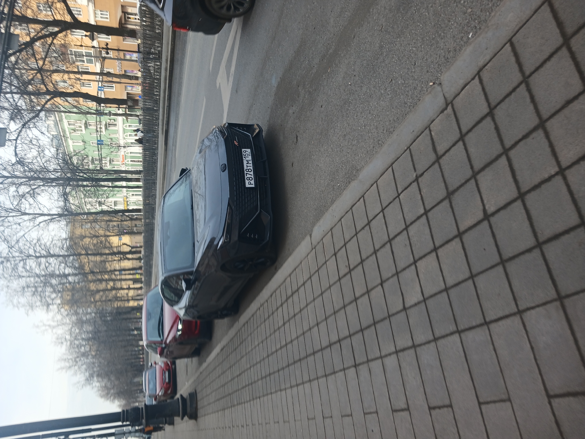 The image shows a street scene in Perm, Russia, with several cars parked along the side of the road. The license plate of the first car is P878TM 59. The street is lined with trees, and there are buildings visible in the background. The sidewalk is made of gray, rectangular paving stones. The weather appears to be overcast.