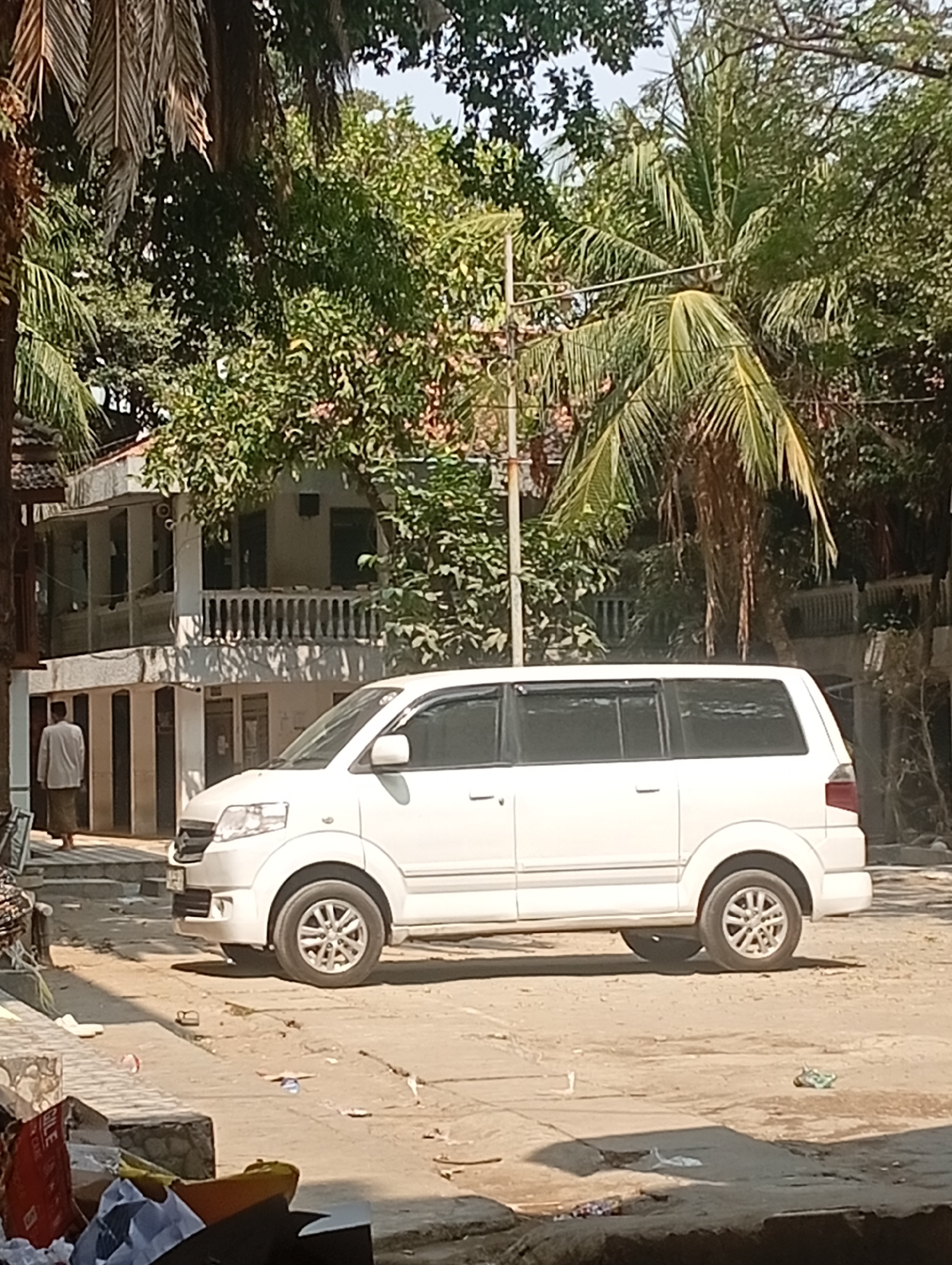 A white minivan is parked on a dirt road in front of a two-story building with balconies. A person wearing a white shirt and sarong is walking away from the building. Lush green trees surround the building and the minivan. The ground is littered with debris, including plastic bottles and papers. The scene appears to be in a residential area or a small town. The location is Bajingjowo, Indonesia.