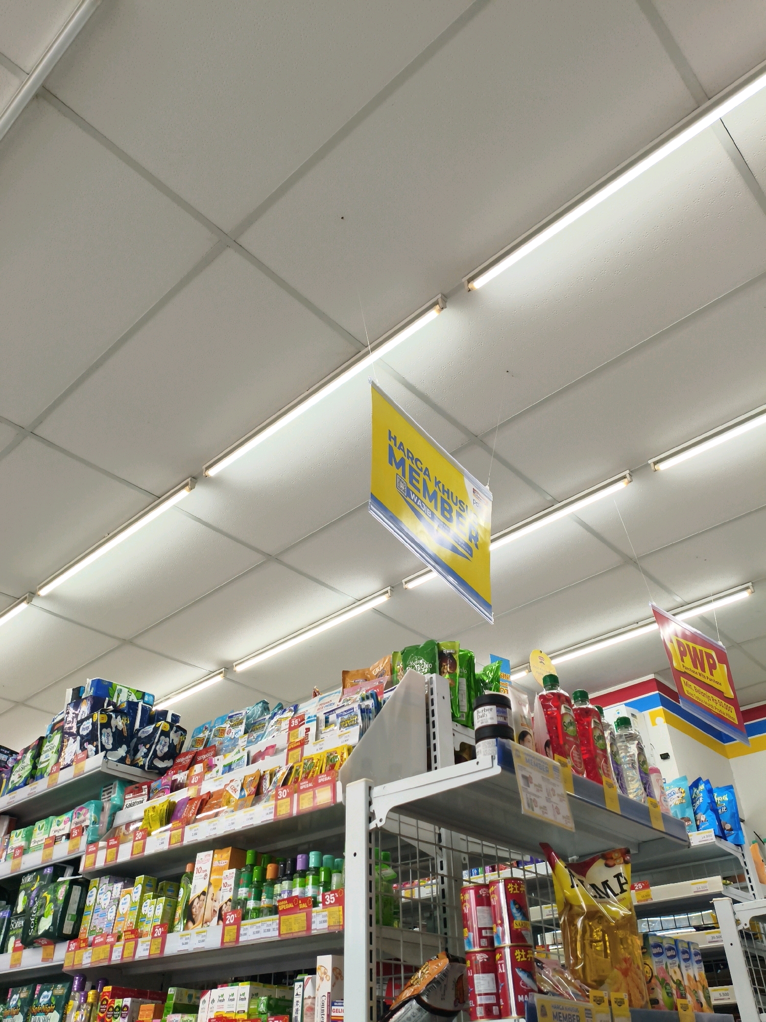 The image shows an indoor view of a convenience store, likely a minimarket, in Sumberejo Kulon, Indonesia. The camera is angled upwards, focusing on the ceiling and the shelves of products. The ceiling is made of white acoustic tiles with fluorescent lighting fixtures. Hanging from the ceiling are two promotional signs. One sign, yellow with blue text, reads "HARGA KHUSUS MEMBER" (Special Price for Members) and "WAJIB" (Mandatory). Another sign, red and yellow, reads "PWP" (Purchase with Purchase) and indicates a minimum purchase requirement of Rp 50,000.
Below the signs, several shelves are stocked with a variety of consumer goods. These include packaged food items, toiletries, cleaning supplies, and beverages. Products are arranged in neat rows, with price tags and promotional stickers visible on many of them. Among the visible products are packs of sanitary napkins, bottled drinks, and various food packages. The overall impression is of a well-organized retail environment designed to attract customers with special offers. No people are visible in the frame.