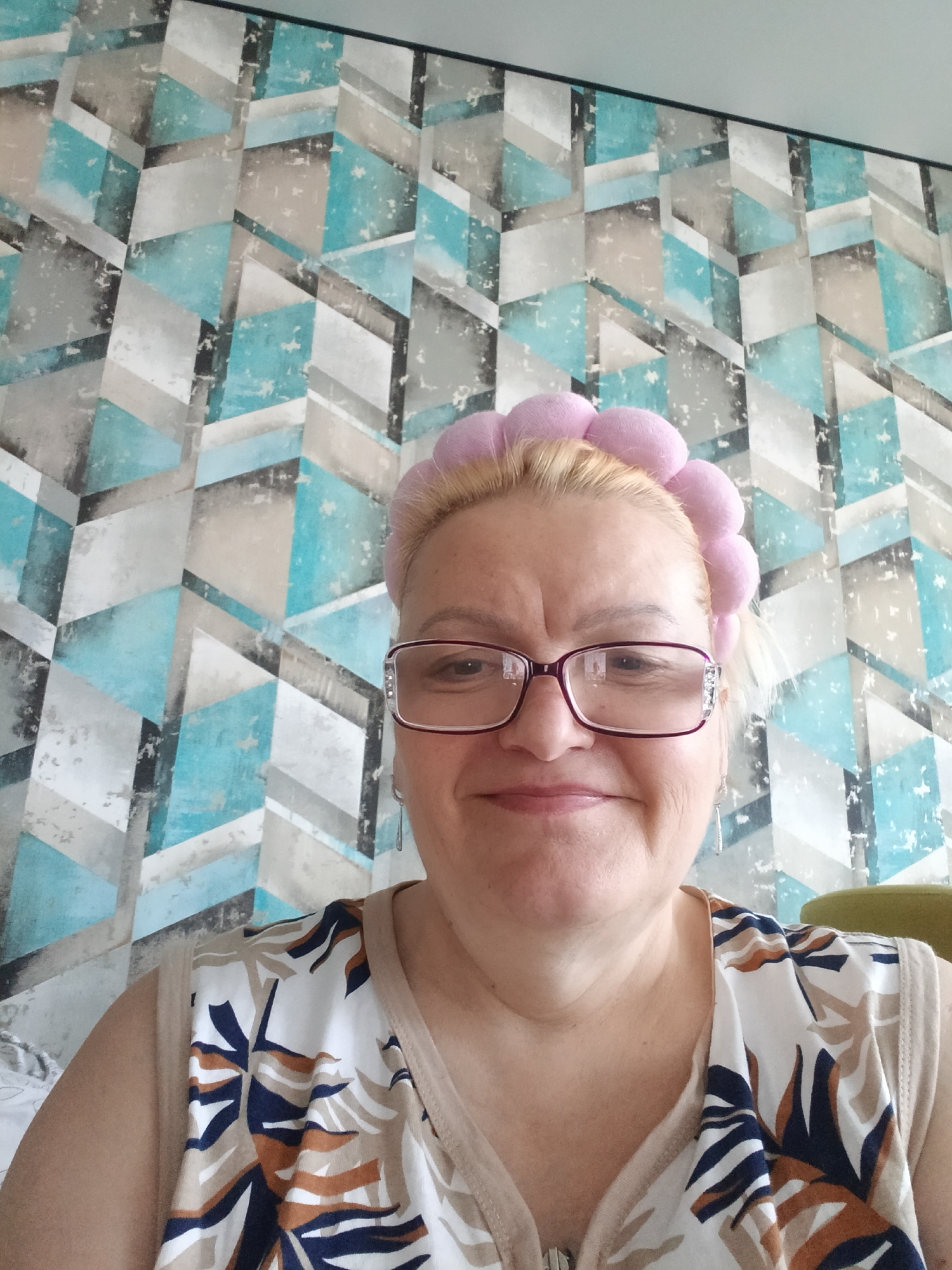 A middle-aged woman with blonde hair and glasses is the subject of this close-up photo. She is wearing a pink headband with round elements and a patterned tank top. The background is a geometrically patterned wallpaper in shades of blue, grey, and white. The photo appears to have been taken indoors. There is no indication that this image was taken in the city of Murom, Russia.
