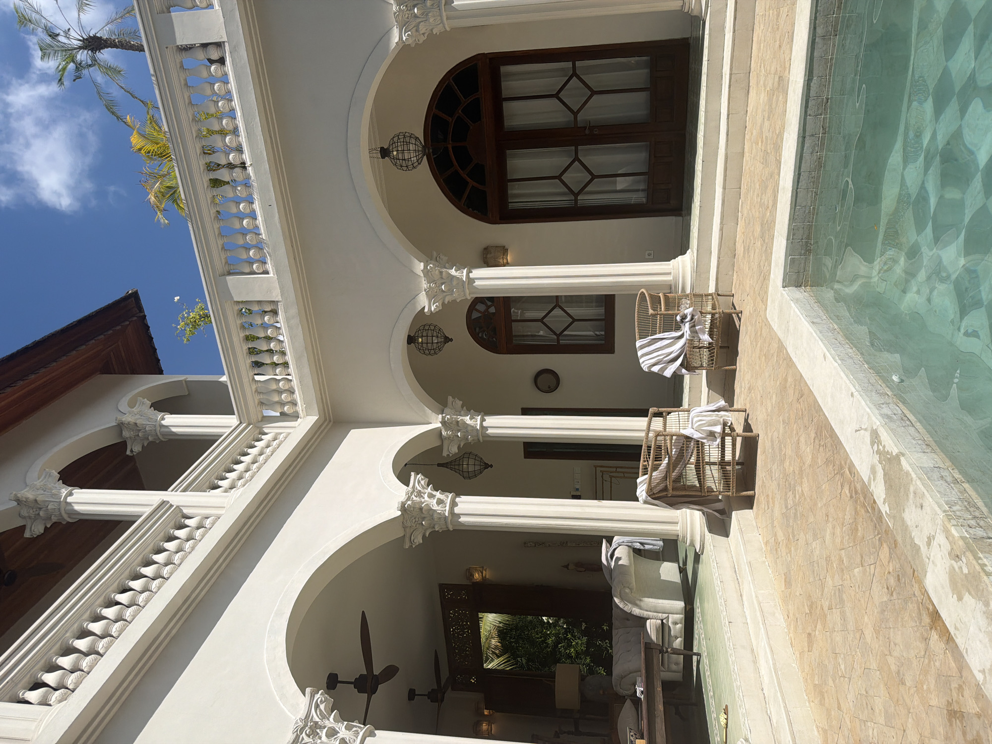 The image displays a luxurious architectural setting, likely a villa or resort, under a bright blue sky with scattered white clouds. The building features an elegant, classical, or Mediterranean-inspired design with pristine white stucco walls, multiple arches, and robust columns topped with ornate capitals. Intricate balustrades, reminiscent of a balcony railing, line the upper levels, adding to the sophisticated aesthetic.
Through the arched openings, glimpses of interior spaces are visible. These include dark wooden doors and windows, some featuring arched tops and geometric pane designs. Ornate spherical cage-like pendant lights hang from the ceilings of these covered areas, alongside visible ceiling fans, indicating a tropical climate. One archway reveals a comfortably furnished indoor living space with a large, upholstered sofa and a table.
On the right side of the frame, a clear blue-green swimming pool shimmers in the sunlight, its checkerboard tile pattern visible beneath the water's surface. Steps lead into the pool, which is flanked by a light brown tiled patio. Two woven rattan chairs, each draped with a striped towel, are positioned on this patio, inviting relaxation. Tall palm trees reach into the bright sky on the left, further emphasizing the tropical environment.
The scene is devoid of people, conveying a sense of peace and tranquility. The bright sunlight suggests it is daytime, with clear weather. The architecture, tropical vegetation, and pool are consistent with a high-end property in a warm climate, such as North Kuta, Indonesia. No visible text is present in the image.