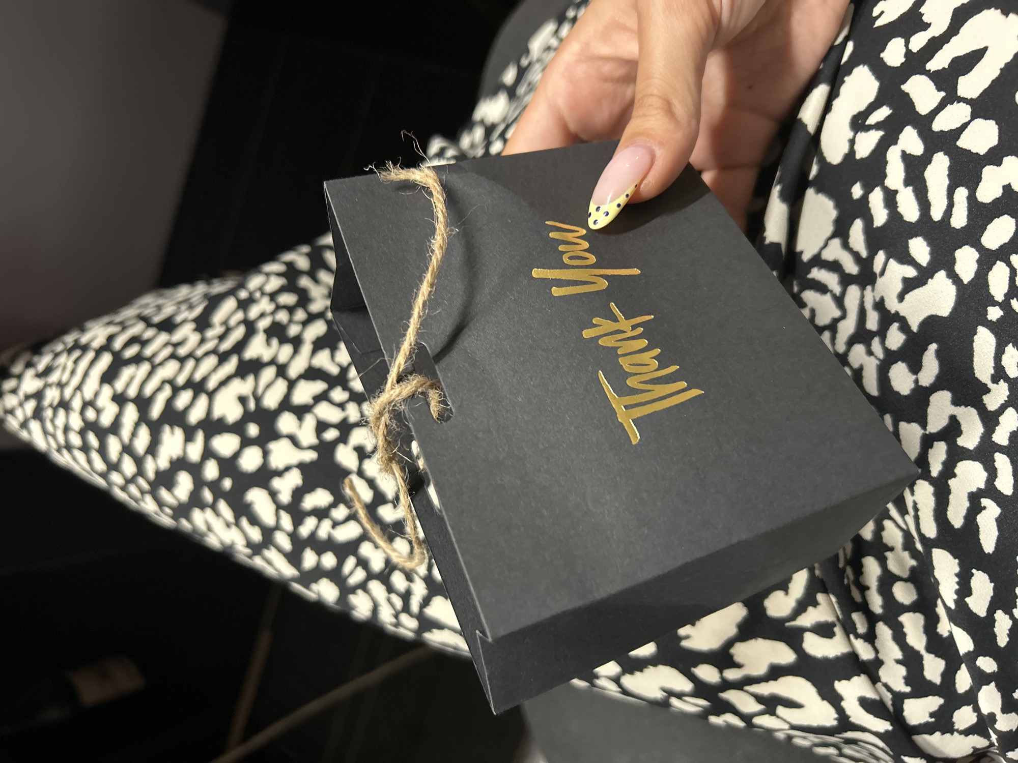 The image shows a close-up view of a black gift box with "Thank You" written in gold foil lettering. A person's hand, with manicured nails featuring a yellow and black polka dot design, is holding the box. The box is tied with a rustic twine string. The person is wearing clothing with a black and white leopard-like print. The background is dark, with some soft lighting, suggesting an indoor setting. The image appears to have been taken in Tbilisi, Georgia. The overall mood is appreciative and elegant.