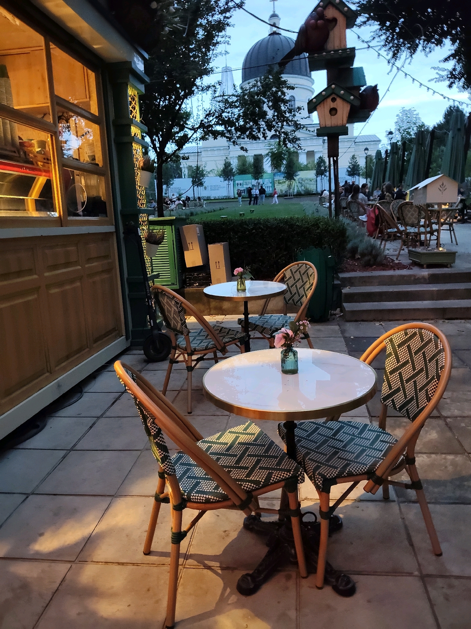 This image depicts an outdoor seating area of a cafe in Chișinău, Moldova, likely in the vicinity of the Nativity Cathedral, visible in the background. In the foreground, there are two round tables with white tabletops and ornate dark bases. One table has a small glass vase with pink flowers. Surrounding the tables are four chairs with woven rattan frames and seat cushions featuring a dark green and white geometric pattern. The chairs are positioned around the tables as if ready for patrons.
To the left, part of a wooden structure, possibly a food stand or cafe counter, is visible. It has large glass windows displaying internal lighting and shelves with stacked items. Adjacent to this structure is a dark green, louvered door and a parked electric scooter.
The background reveals a park-like setting with a manicured lawn, trees, and pathways. Several groups of people are seated at tables in the distance, and others are standing and walking. A distinctive tiered birdhouse structure with bird figurines stands prominently on a pole. Further in the background, the domed Nativity Cathedral and other buildings are silhouetted against the twilight sky, suggesting the photo was taken in the early evening. The overall atmosphere is that of a relaxed outdoor dining or gathering space in a city park.