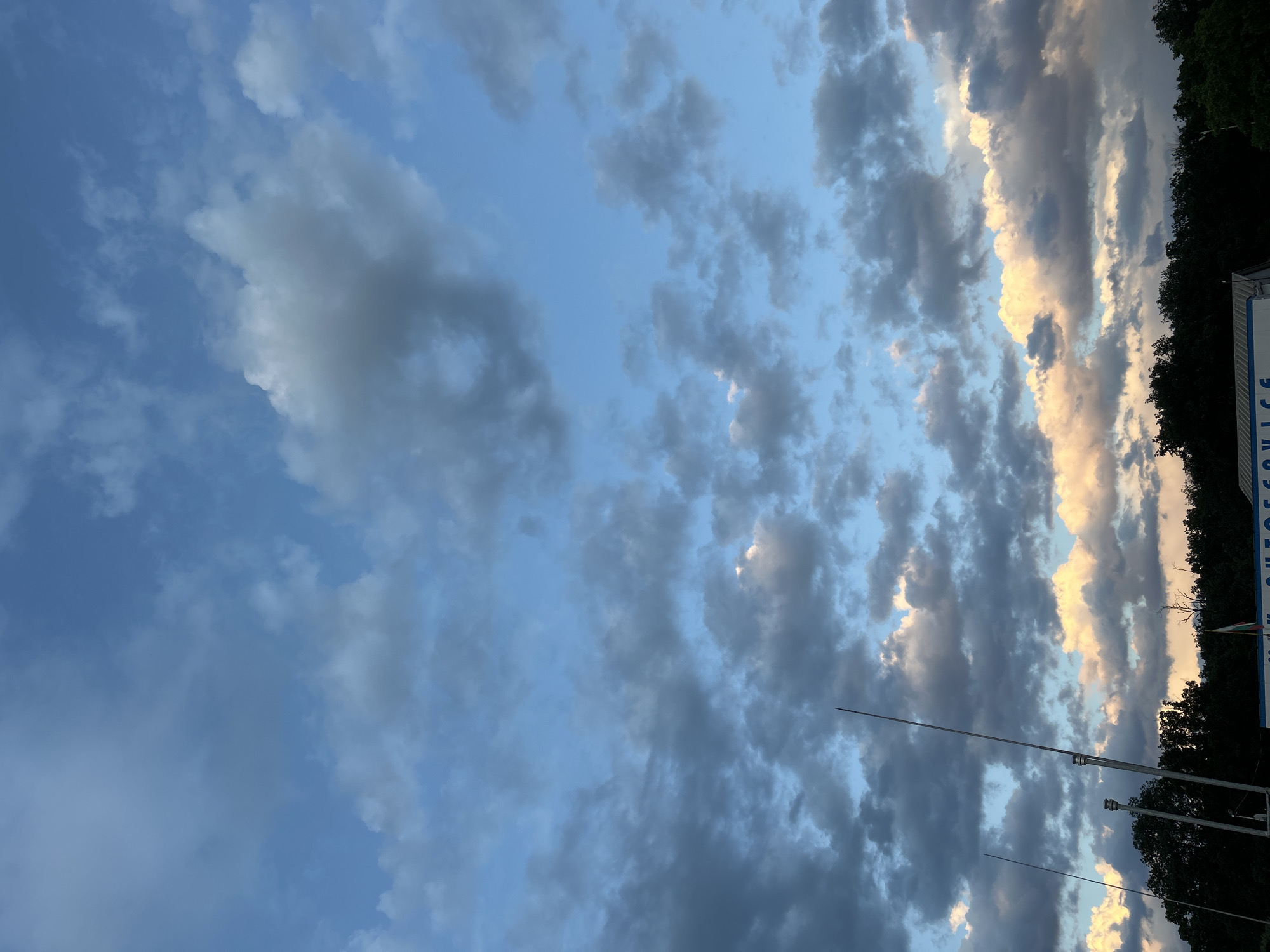 The image captures a dramatic sky filled with a variety of cloud formations during what appears to be either sunrise or sunset. The sky is predominantly covered by clouds, ranging from dark grey, dense clusters to softer, brighter white and golden-orange illuminated sections. Patches of clear blue sky are visible in between the cloud masses.
Towards the bottom right of the frame, a dark silhouette of a tree line or dense foliage is visible. Below this, a structure or a large sign is partially seen, featuring the legible text "AUTOSERVICE" in blue letters on a white background. Next to the "AUTOSERVICE" sign, a small flag or emblem with visible colors, potentially resembling the Bulgarian flag, is present. Two thin, tall poles, possibly antennas or flagpoles, extend upwards from the bottom of the image, reaching into the cloudy sky.
The overall scene, taken in Krepost, Bulgaria, conveys a serene yet visually striking atmosphere, characterized by the interplay of natural light on the diverse cloud structures and the subtle presence of man-made elements at the horizon. The warm tones on some clouds suggest the soft, directional light of either the early morning or late afternoon sun.