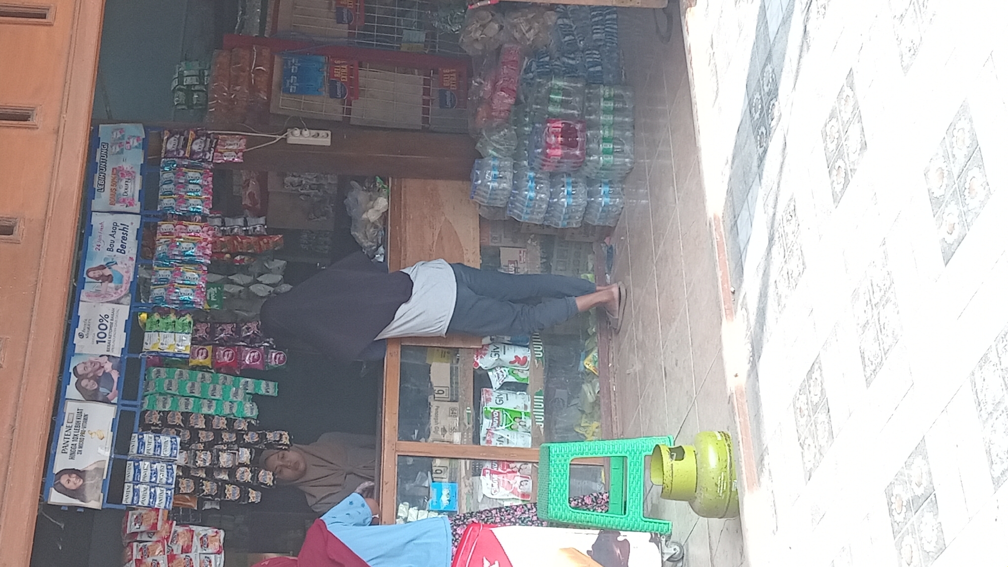 A street-level view captures a small retail establishment in Kedungwaru, Indonesia, likely a warung or minimarket, where goods are displayed both indoors and outdoors. The facade is a wooden structure with a prominent overhang. Inside, shelves are densely packed with a variety of packaged consumer goods, including sachets of snacks, hygiene products like soap, and bottles of beverages. Two individuals are visible behind the counter. One, wearing a light brown hijab and a light-colored top, appears to be serving or attending to a customer. Another individual, wearing a dark hijab and a light grey and dark blue top, stands with their back to the camera, interacting with the establishment's interior. Outside the store, stacks of bottled water are arranged on the ground to the right, and a green gas cylinder sits to the left, next to a green plastic stool. The floor is tiled with a mosaic pattern. Sunlight casts strong shadows across the scene, indicating it is daytime.
