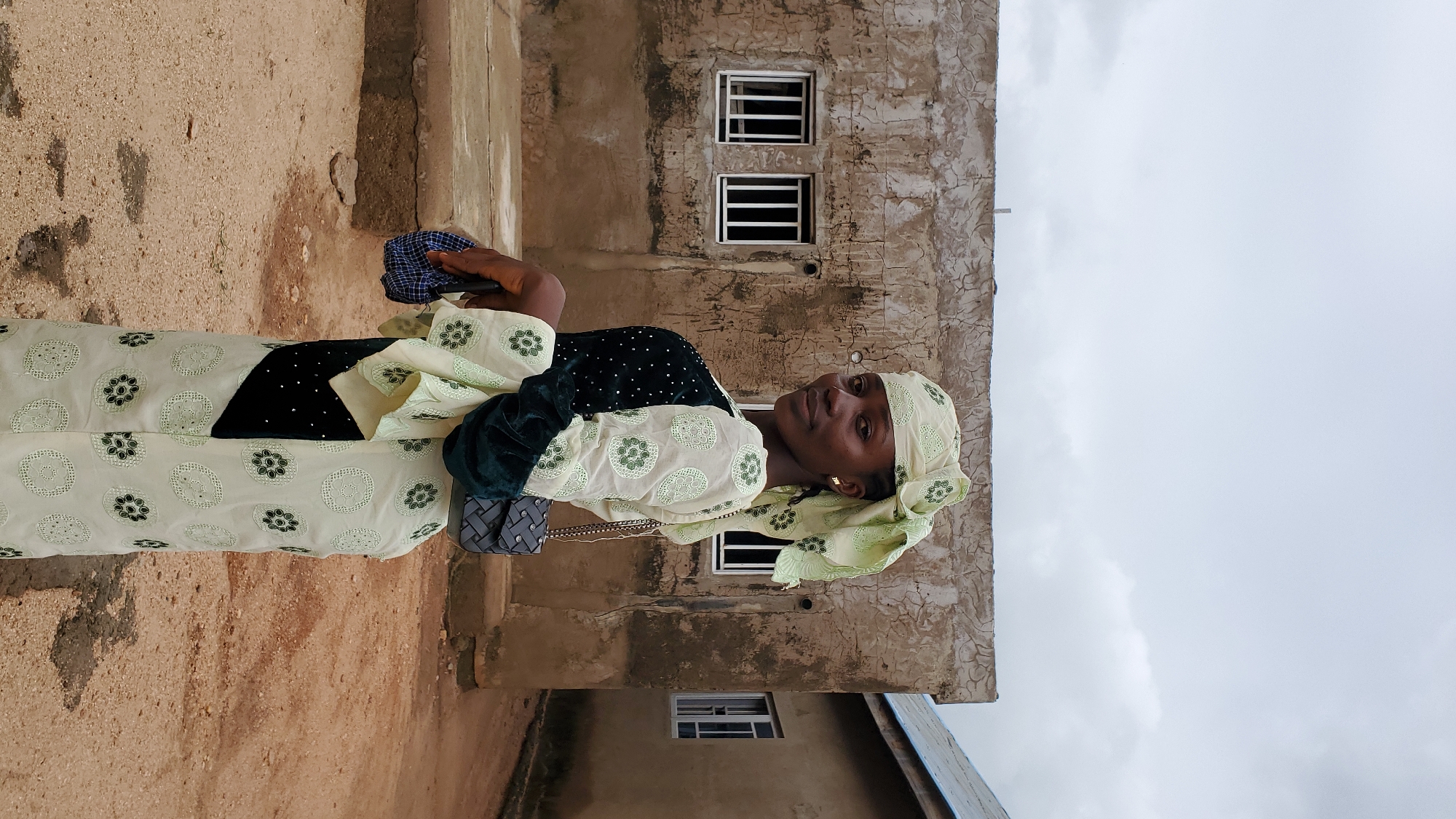 A young woman stands outdoors in front of a textured, pale brown building with barred windows. She is wearing a light green and cream patterned outfit with a matching headscarf. A dark green velvet trim accents the sleeves of her garment. She is carrying a small, dark grey handbag with a chain strap over her shoulder and holding a folded blue and white checkered object in her left hand. The ground in front of her is dirt. To the right, another building with a light blue roof is partially visible. The sky is overcast and pale grey. This scene is likely located in the city of Jalingo, Nigeria.