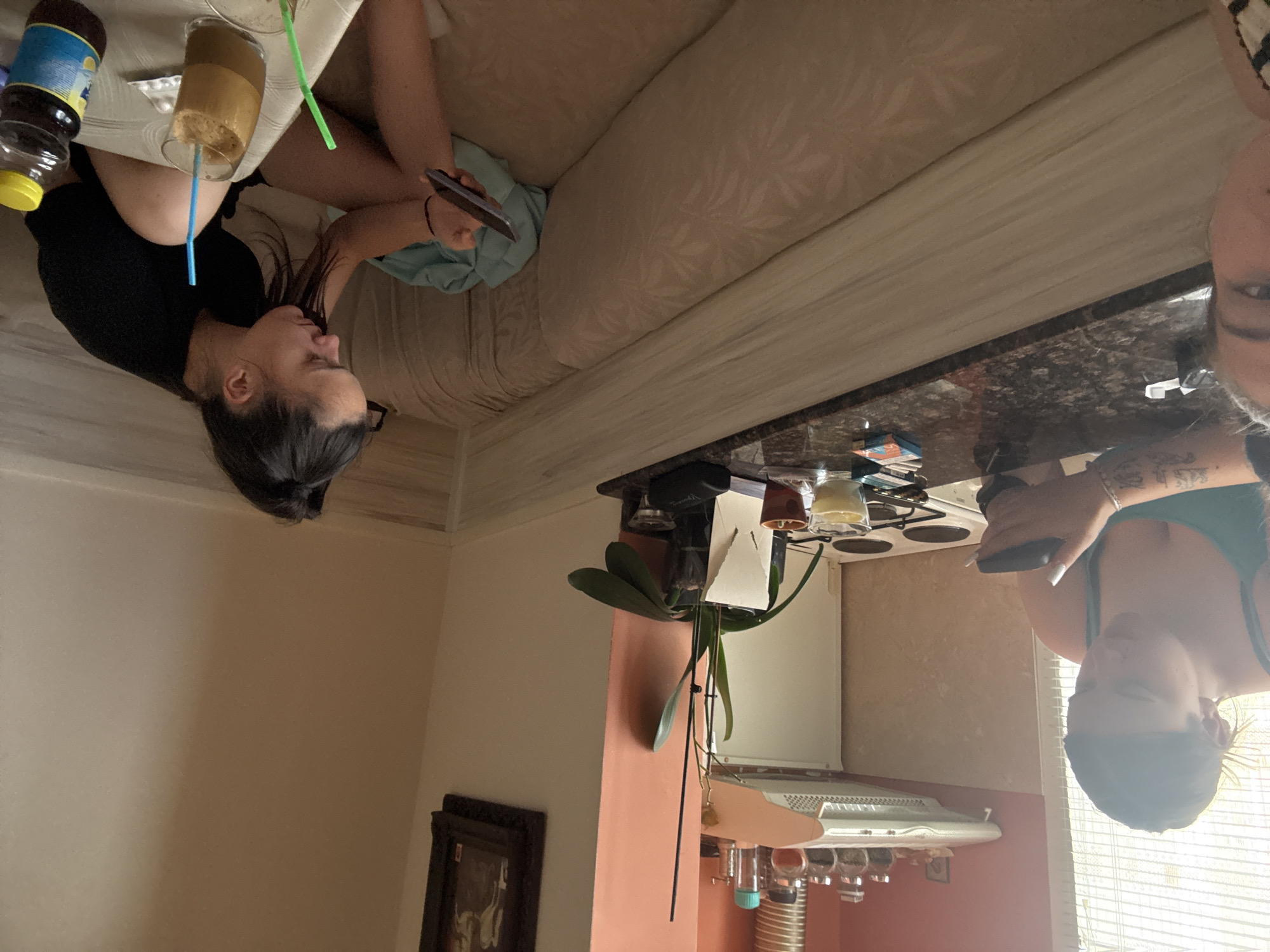 The image, rotated 90 degrees clockwise, depicts an indoor scene in what appears to be a home in Kyustendil, Bulgaria. Two women are visible, engaged in relaxing activities, likely during the daytime.
In the foreground, a woman with dark hair and glasses, wearing a black top, is seated. She is looking down at a smartphone held in her left hand. On a surface beside her, there is a tall glass containing an iced coffee or frappe with both a blue and a green straw. Next to the drink, a clear bottle with a yellow cap and a blue label is visible, bearing the text "LEMON" and what appears to be "LIMONADE" or "KABANA." A beige couch with a subtle floral pattern extends behind her.
Further into the background, another woman with dark hair, wearing a green top, is seen from the chest up. She is also looking down at a smartphone. Her right arm displays tattoos, the details of which are not clearly legible. She appears to be in a kitchen area, with a dark, speckled countertop and a stovetop visible behind her. A potted plant, possibly an orchid, sits on a shelf above the counter. Below the counter, kitchen fixtures including a range hood and hanging cups are present. Natural light enters the room through a window with blinds on the right side of the image. A framed picture hangs on a wall in the bottom left portion of the image. A partial view of a third person's head/face is discernible in the top right corner, looking towards the camera. Both visible women exhibit a relaxed posture, focused on their phones.