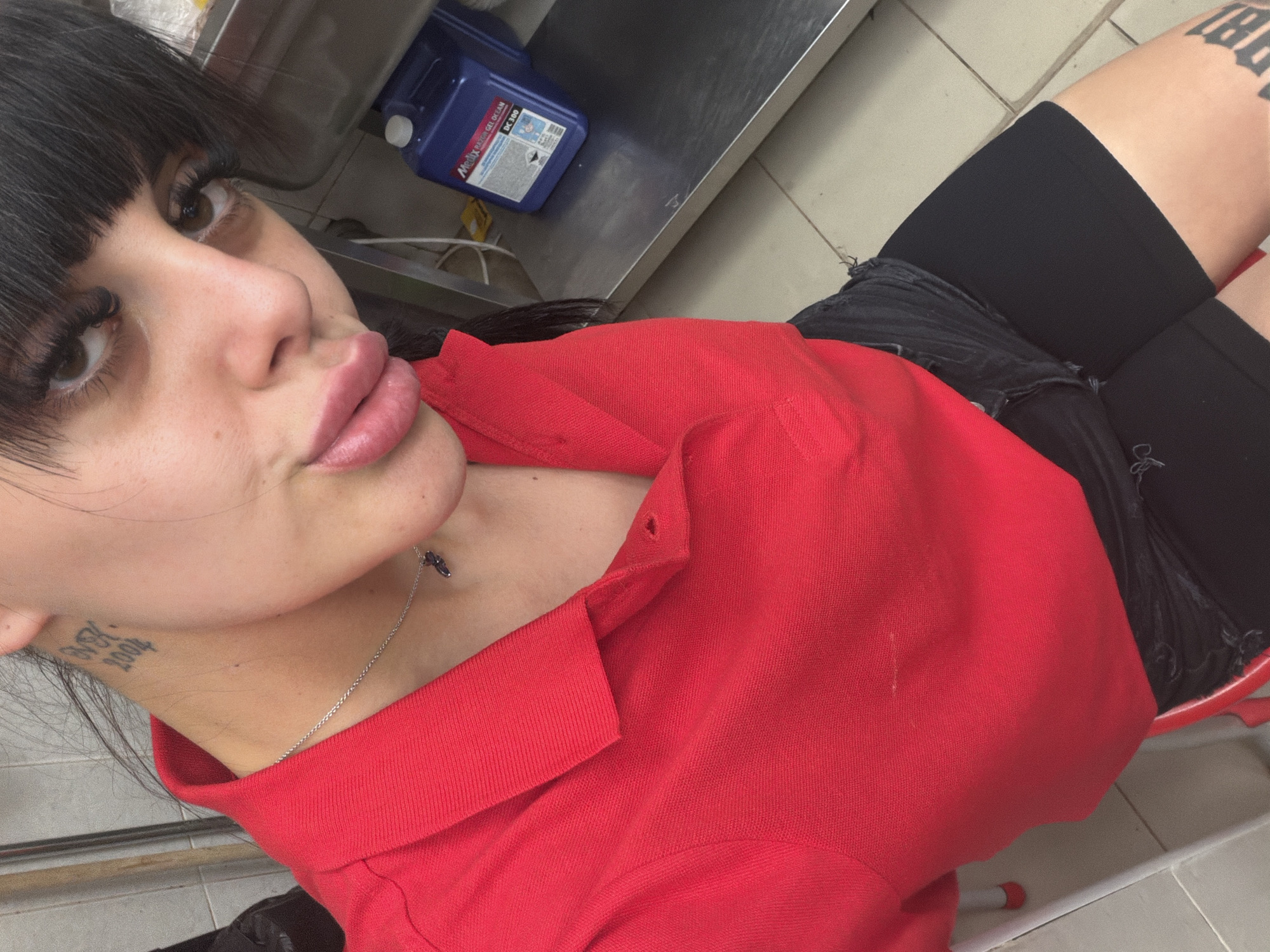The image shows a close-up, high-angle shot of a young woman with dark hair and bangs, wearing a red collared shirt and black shorts. She is looking at the camera with her lips slightly parted. Her eyes are dark brown and her eyelashes are long and full. She is wearing a thin silver necklace with a small pendant. There are tattoos visible on her neck and her upper thigh.
In the background, a blue plastic container with white lettering is visible on a metal shelf. The setting appears to be an indoor environment, possibly a kitchen or a commercial space, given the stainless steel surfaces and tiled floor. The lighting suggests it is daytime. The woman's expression is neutral, but her pose and gaze create a direct connection with the viewer.
Visible text on the blue container includes "MEDIX RACING GEL OCIAM" and "DC 300." There is also text on the woman's thigh tattoo, but it is partially obscured and difficult to read.