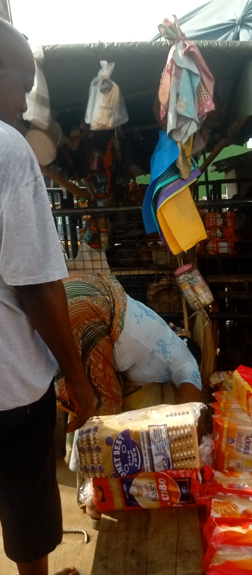 The image depicts an outdoor commercial scene, possibly in Nkpor, Nigeria, centered on a roadside stand laden with various loaves of bread. A person in a patterned skirt and a light blue blouse bends over the bread. A man in a gray T-shirt and black shorts stands at the left. The bread is stacked and wrapped in plastic; brands include "Honey Best Rich Food" and "UBO." Hanging above the bread are various objects: plastic bags with small items, cloth, and containers. The structure sheltering the stand appears to be a makeshift arrangement of wood and fabric. The overall setting suggests a busy commercial area with people engaged in transactions.
