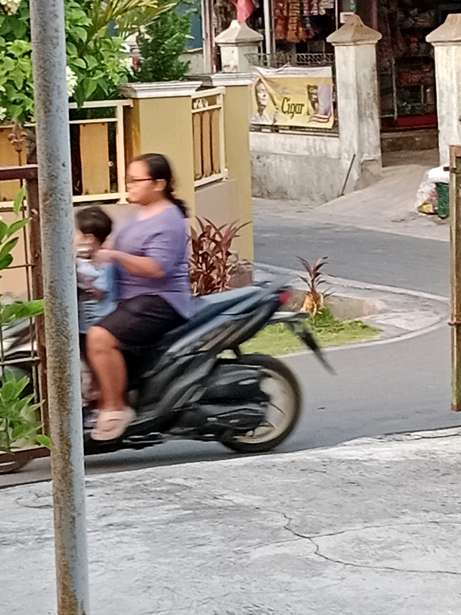 The image shows a woman wearing a purple shirt and a dark skirt riding a dark-colored scooter. She has glasses and her hair is pulled back. A child wearing a light blue shirt is seated in front of her on the scooter, facing the woman. They are riding on a paved road next to a sidewalk and a low wall with decorative elements. In the background, there is a building with a yellow facade and a banner that reads "Cigar" with a picture of a face. Green plants are visible on the left side of the image. The overall environment appears to be a street in an urban or semi-urban area, possibly in Waru, Indonesia.