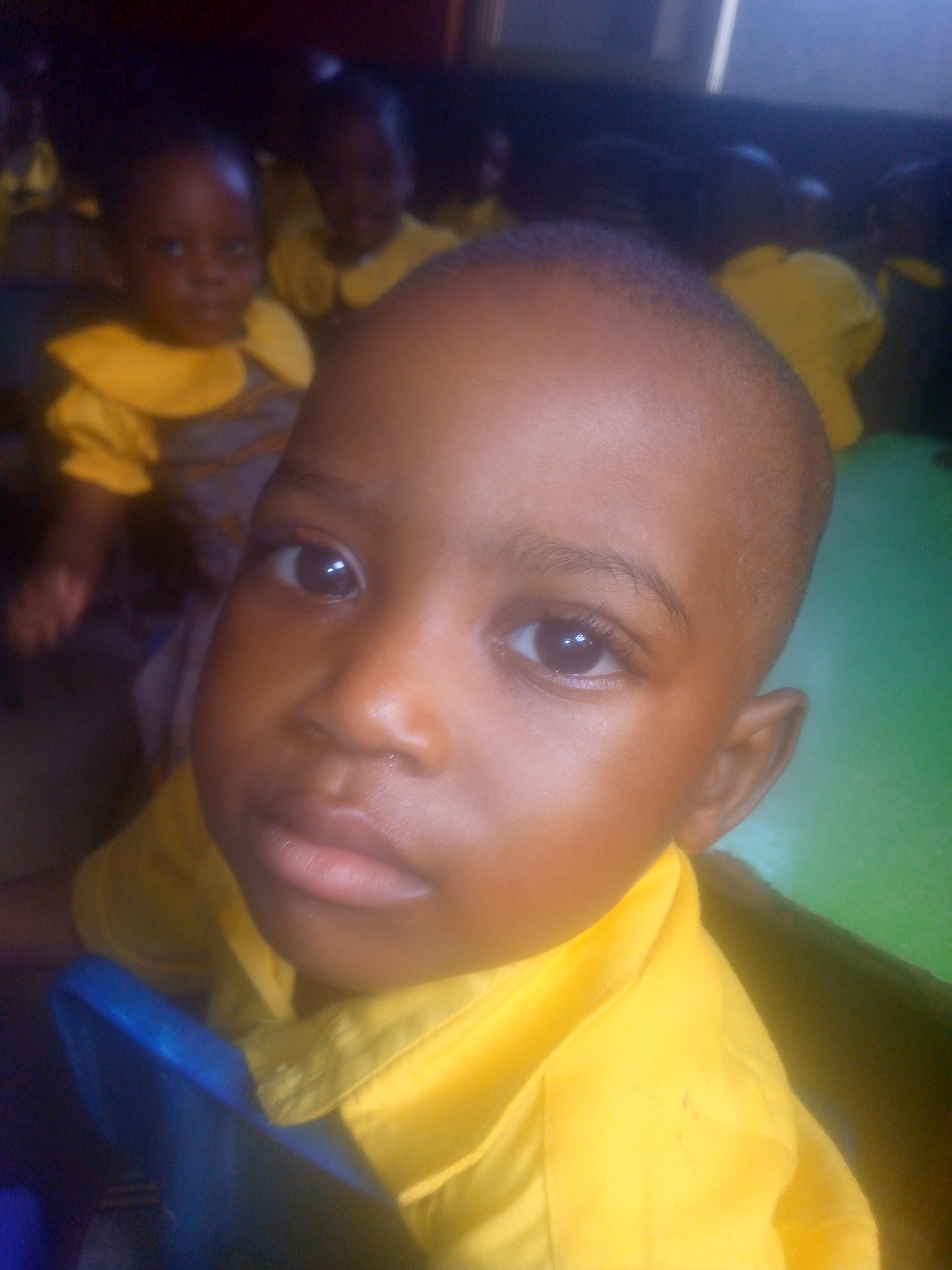 A young child with dark skin, likely a boy, is the primary subject, appearing in a close-up shot. He has short, neatly trimmed hair, and wears a bright yellow shirt. His expression is neutral, his eyes gazing directly into the camera.
Several other children, also with dark skin, are visible in the background. They wear similar yellow and gray outfits, possibly uniforms, suggesting a school setting.
The location is unspecified, but the context suggests it could be a school or classroom in Ikorodu, Nigeria. The environment includes some green surfaces, possibly tables or walls.
The primary action is the child posing for the photo. The interaction is between the child and the photographer.