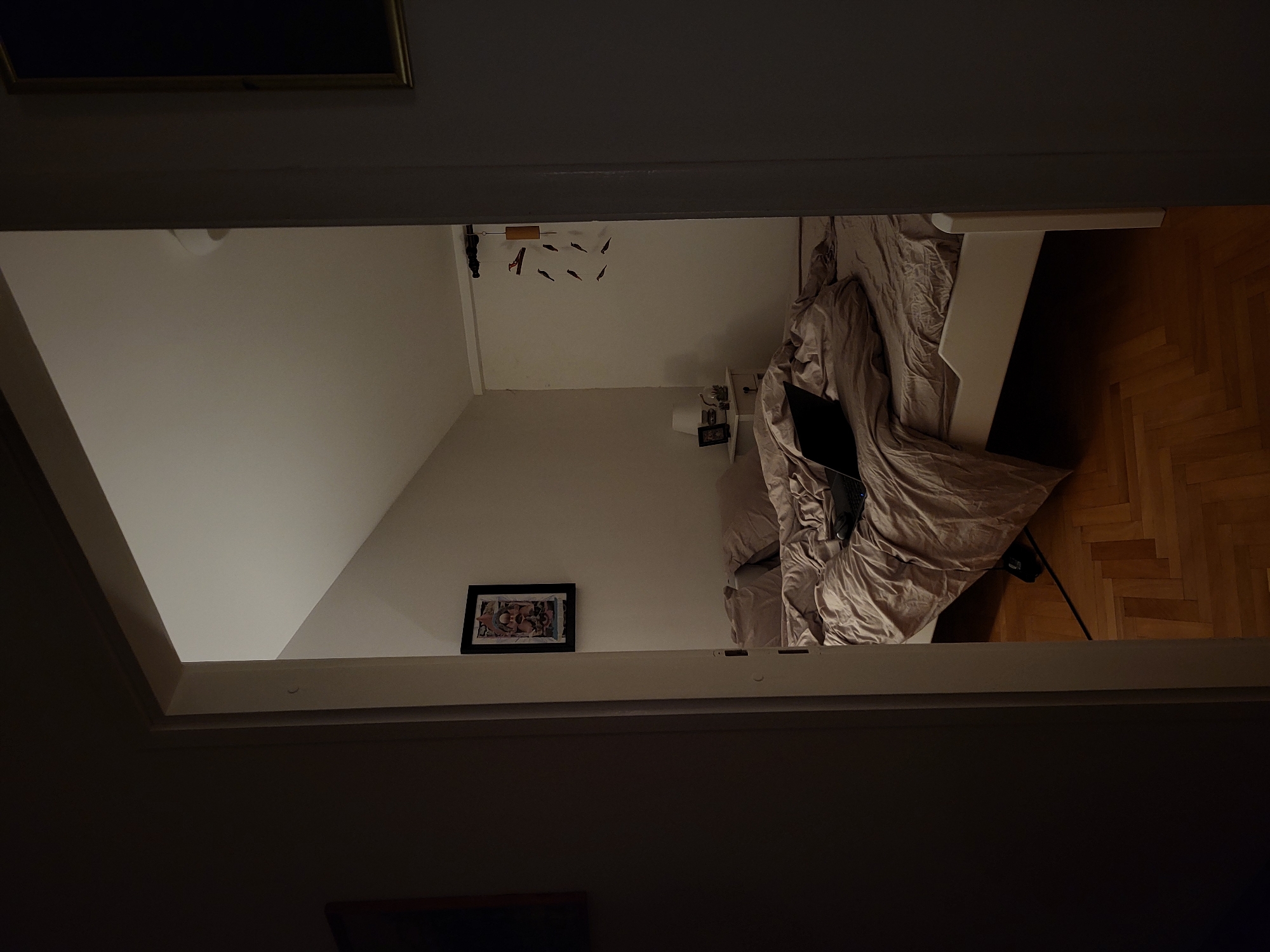The image shows a bedroom viewed through an opening, possibly a loft access or a doorway. The room contains a bed with rumpled beige-colored bedding, a laptop, a nightstand with decorative items, and a framed picture hanging on the wall. The flooring is herringbone-patterned wood. Above the bed, a mobile with several small figures hangs from the ceiling. The perspective is from above, looking down into the room. The lighting is soft and dim, with the bedroom being brighter than the foreground area where the camera is positioned.