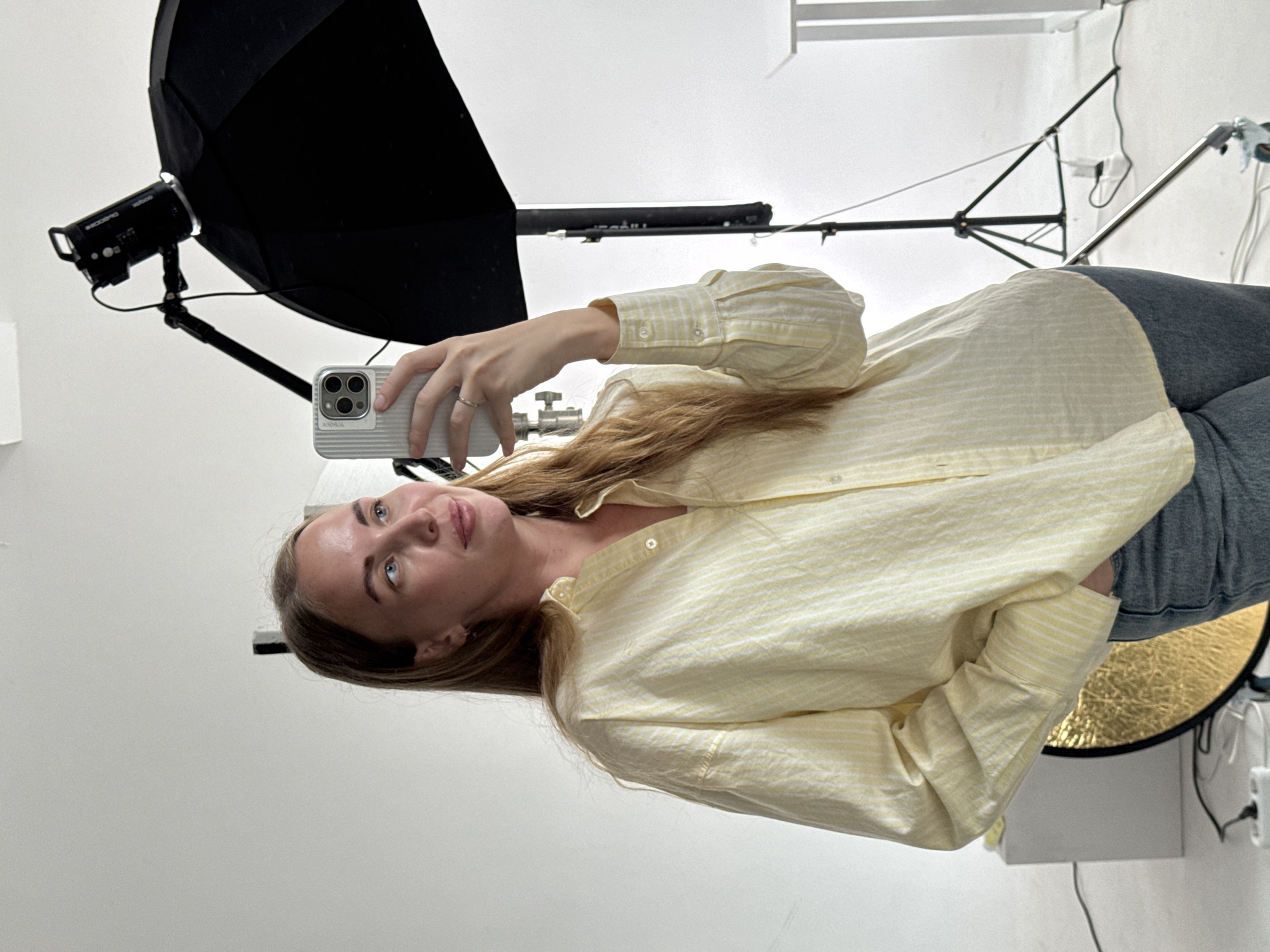 A fair-skinned woman with long, light brown hair and blue eyes is taking a selfie with a white iPhone. She is wearing a yellow and white striped long-sleeved shirt and gray jeans. She is standing in what appears to be a photography studio, with lighting equipment including a softbox and a reflector visible. The background is plain white. There is no indication of this image being taken in the city of Sochi, Russia.