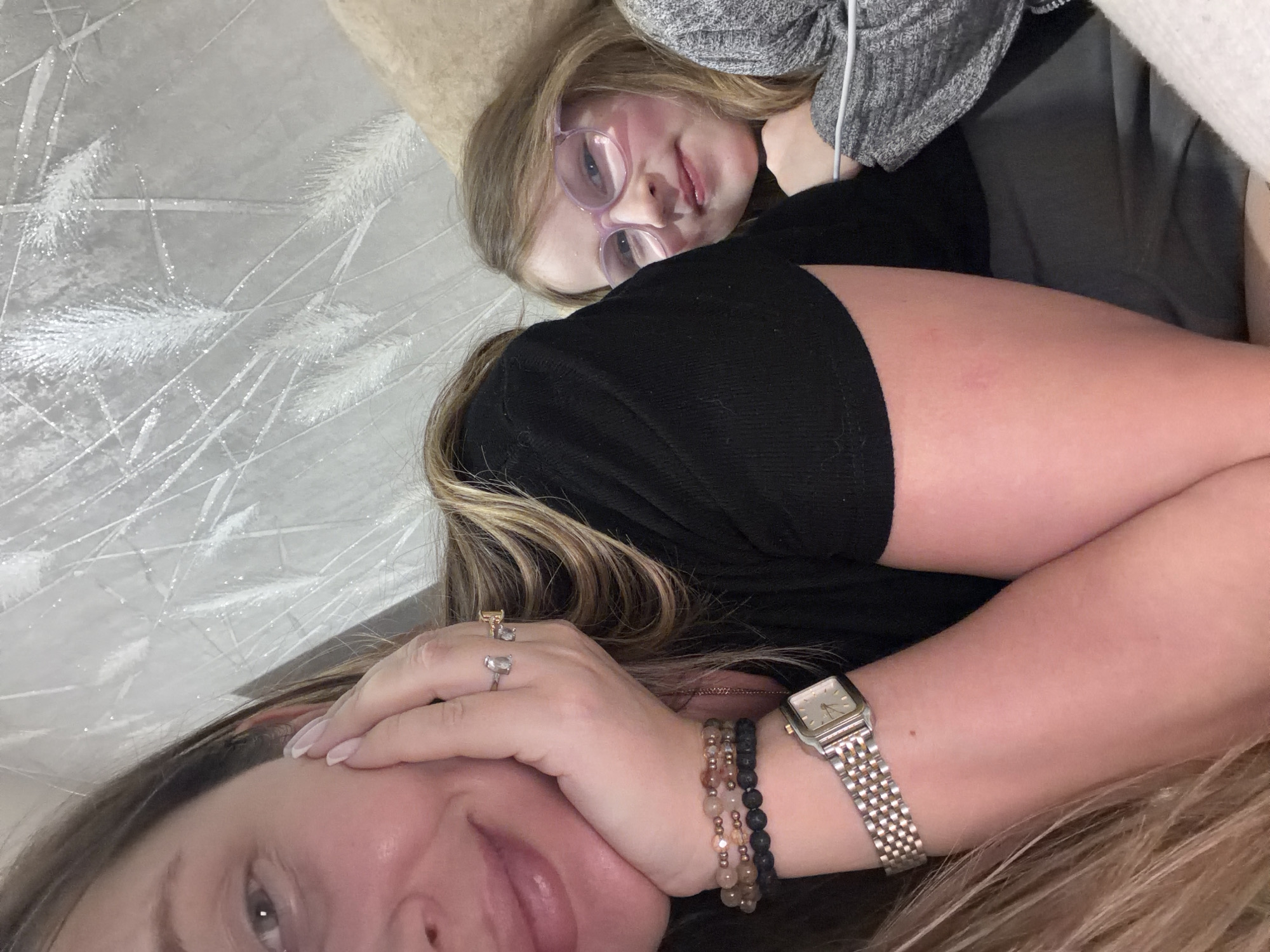A selfie-style photograph shows two individuals lying down. At the top of the frame, a young girl with blonde hair and pink glasses is angled towards the camera. She is wearing a grey knit top. Below her, an adult woman is lying down, facing the camera with her left hand near her face. She has blonde hair and is wearing bracelets and rings. A watch is visible on her wrist. The background appears to be a textured surface with a white, decorative pattern. There is no definitive evidence to confirm the location as Chaikovsky, Russia.
