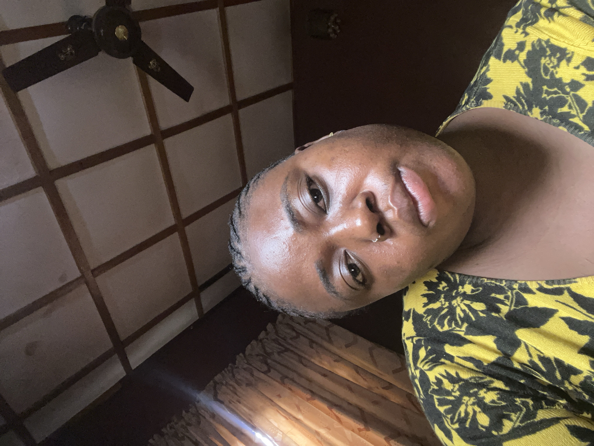 A person is seen indoors, lying on their back, facing the camera. The person is wearing a yellow top with dark floral designs. The background includes a ceiling with a dark ceiling fan and a grid-like pattern. A curtain is partially visible. There's no information to confirm the image was taken in Jos, Nigeria.