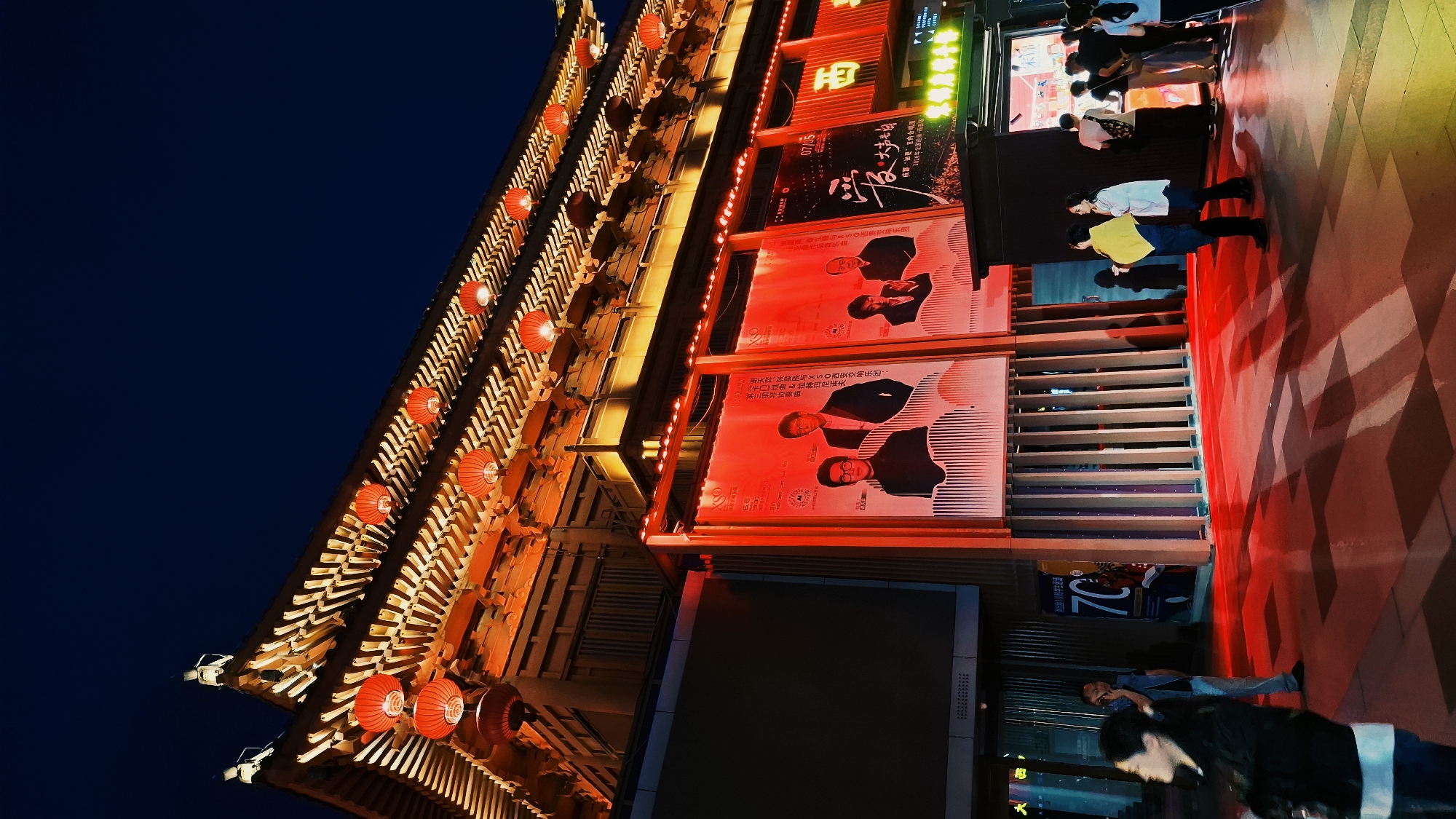 A night shot depicts a brightly lit, traditional Chinese building in Yanta District, China. Red lanterns adorn the multi-tiered roofline of the building, which features ornate wooden architecture. The building's facade is covered in large posters, illuminated with red light. People are walking by, some looking at the posters, and their silhouettes are visible against the bright backdrop. A dark blue sky is visible above the building. The ground is also illuminated with red lighting.