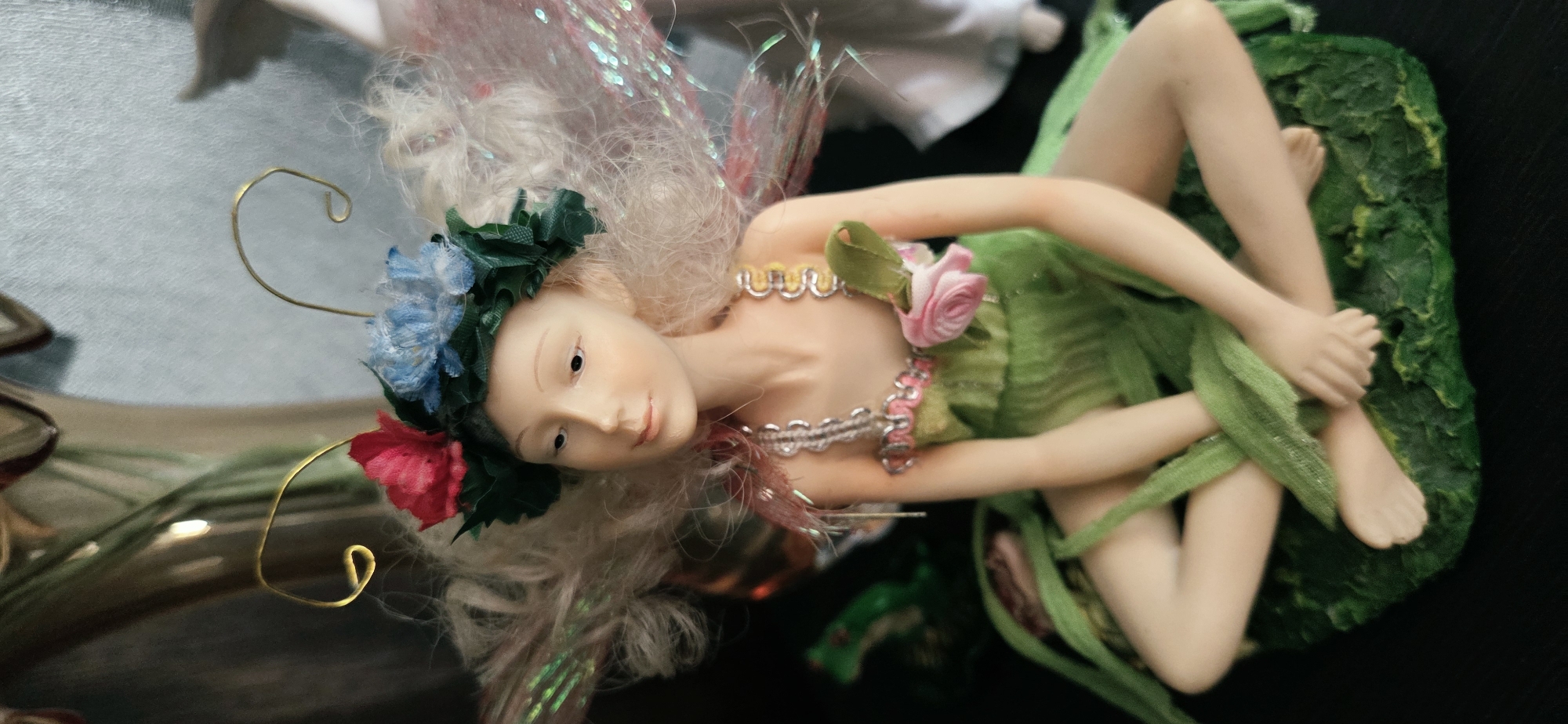 This image depicts a close-up, eye-level, vertical shot of a decorative fairy figurine seated on a textured green base. The figurine has pale skin, light blonde hair, and is adorned with a floral wreath on its head featuring red and blue flowers. It wears a green dress with pink floral accents and silver chain-like straps. The figurine has delicate, translucent wings with pink and iridescent shimmer. The background is dark and out of focus, with hints of other decorative elements. There is no discernible location in the city of Lipetsk, Russia, as this is a staged indoor scene.
