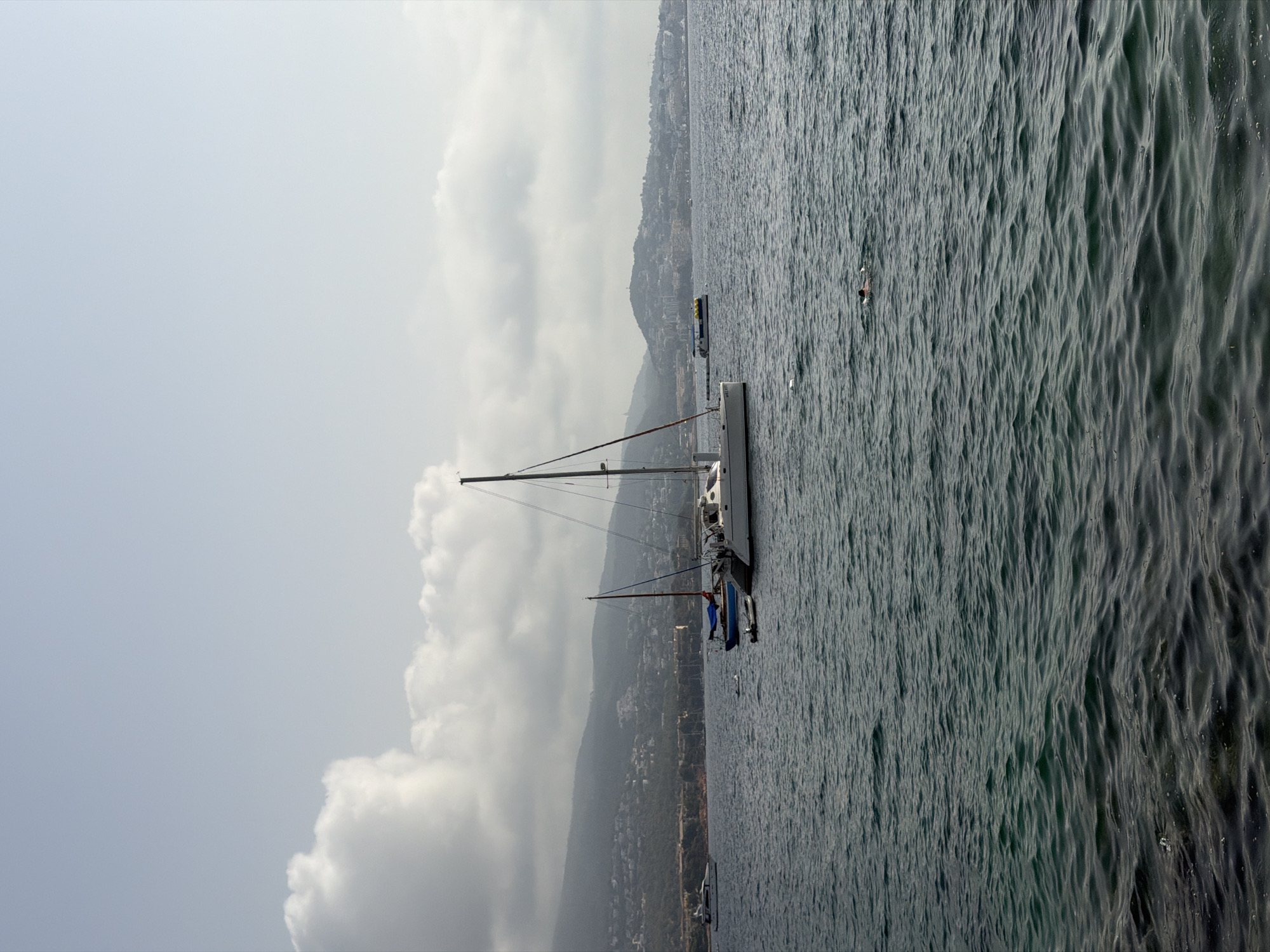 The image depicts a panoramic view of the sea off the coast of Magaluf, Spain. In the foreground, the water is a deep blue-green with visible ripples and small waves. Further out, several boats are anchored. A prominent white catamaran is positioned centrally, with its mast reaching towards the sky. To its left, a smaller blue boat is moored. In the distance, a hazy coastline is visible, characterized by rolling hills covered in scattered buildings and vegetation, indicative of urban development. The sky is a pale blue with scattered, large, white cumulus clouds that cast soft shadows on the landscape. A solitary swimmer is visible in the middle-right portion of the sea, their body partially submerged.