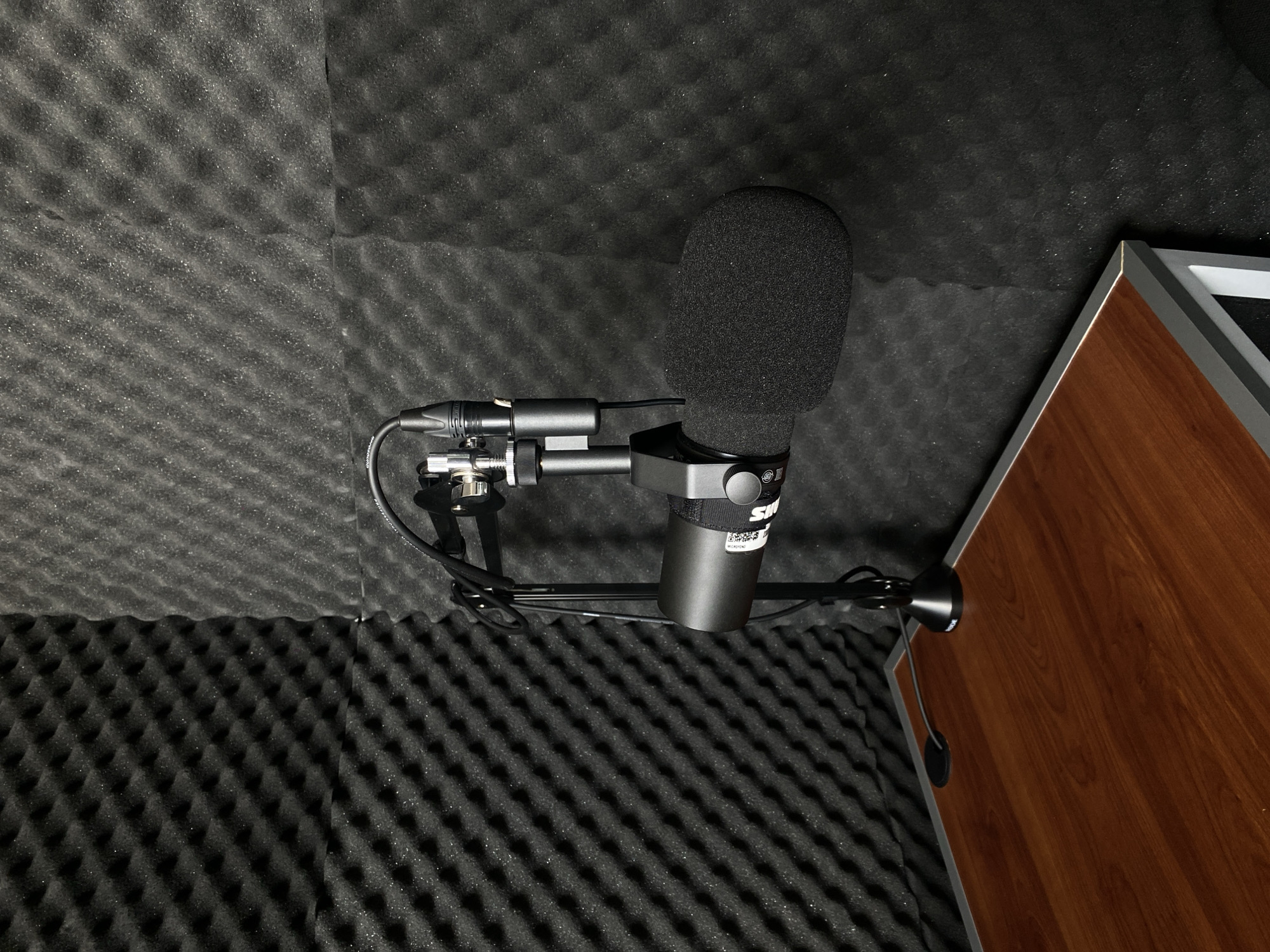 A close-up, overhead shot captures a Shure MV7 microphone inside a room, likely a recording booth, lined with sound-dampening foam tiles. The microphone is mounted on a black stand, complete with a pop filter, and is connected to an XLR cable. Part of a wooden table with a metal edge is visible in the lower right corner. The image gives the impression of a dedicated recording space. No people are visible. The location is Unknown, Unknown.