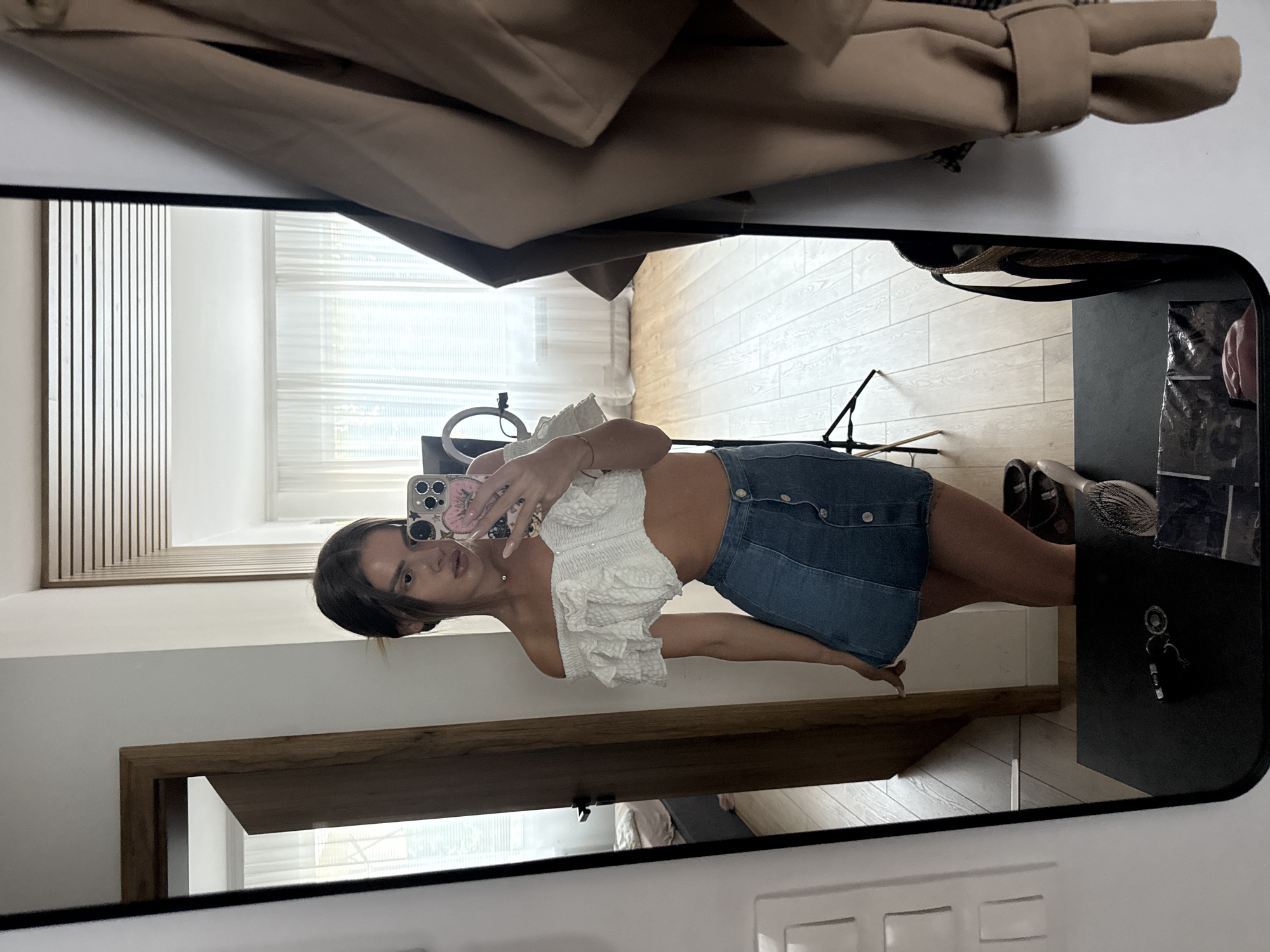 A young woman is taking a mirror selfie in an indoor setting. She is wearing a white off-the-shoulder ruffled crop top and a blue denim mini skirt with a button-down front. The mirror reflects her and the room behind her, which appears to be a bedroom or living area with light-colored walls, wooden floors, and sheer white curtains covering a window. To her left, a doorway reveals a glimpse of another room with a bed. On a table in front of the mirror are keys, a hairbrush, and a basket. A beige jacket is draped over the right side of the mirror. The image does not provide any information to indicate it is located in Plovdiv, Bulgaria.