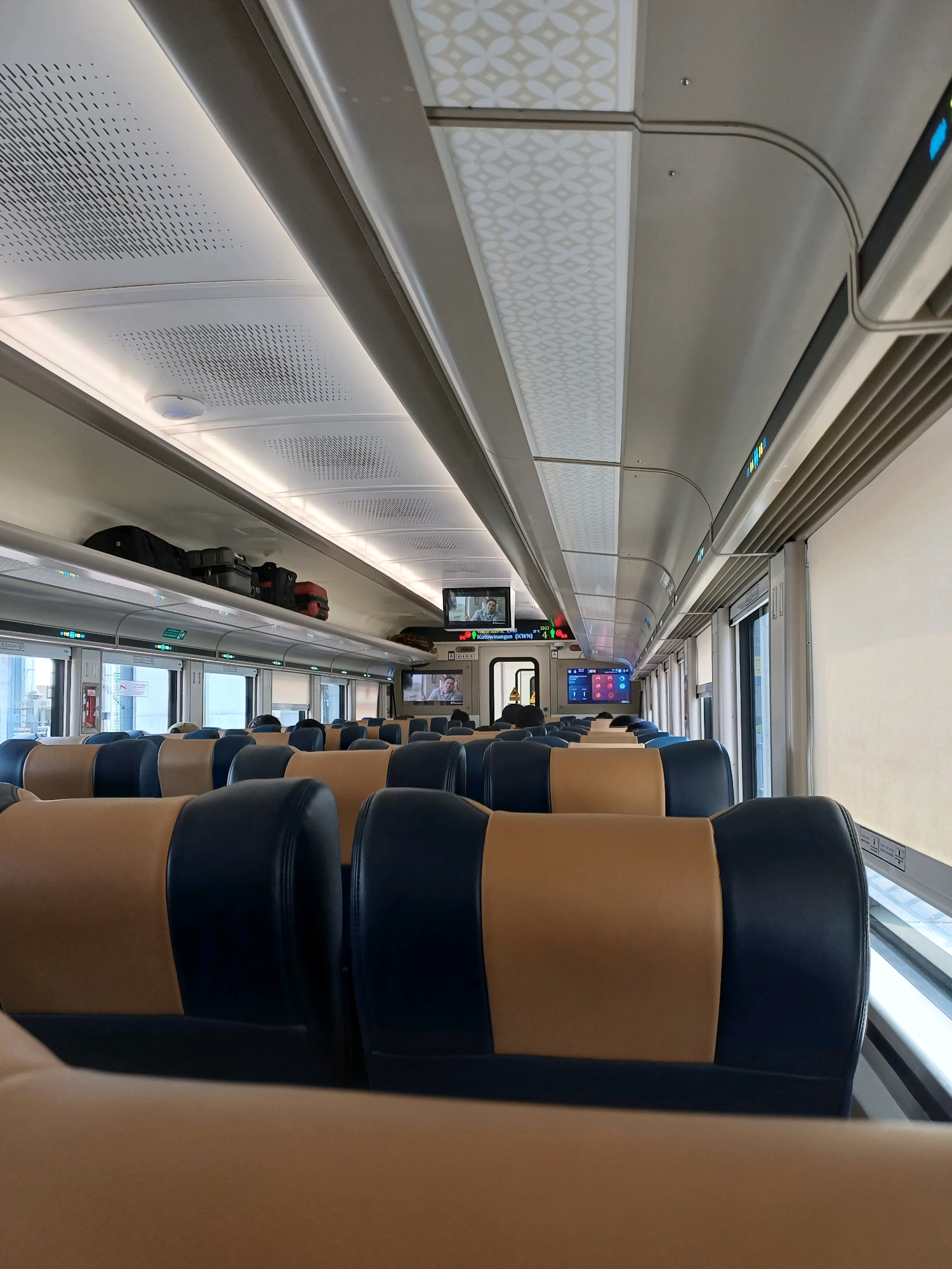 The image depicts the interior of a modern train carriage, likely a commuter or intercity train, as suggested by the presence of rows of seats and overhead luggage racks. The seats are upholstered in a two-tone design, with dark blue on the sides and a tan or light brown color in the center. The carriage features a clean, contemporary aesthetic with a white ceiling adorned with patterned panels and integrated lighting. Several digital screens are visible, displaying route information and possibly entertainment. Luggage is stored in the overhead compartments, suggesting ongoing travel. The windows are covered with light-colored blinds. The train is stationary, as indicated by the lack of motion blur and the orderly arrangement of the interior. The overall impression is one of a well-maintained and comfortable passenger vehicle. While the prompt asks for the location in Surakarta, Indonesia, there are no visual cues in the image itself that definitively confirm this specific location.