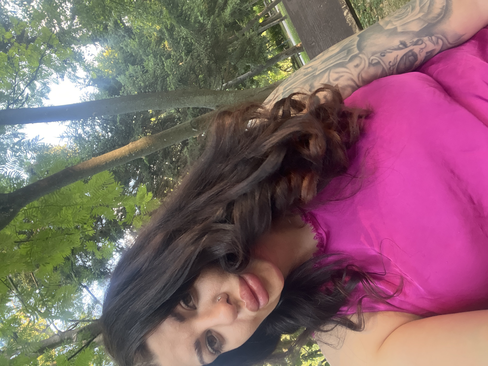 A selfie, taken outdoors, shows a woman with dark, wavy hair and prominent lips, wearing a bright pink top. Her left arm, visible on the right side of the frame, is covered in a detailed tattoo of a skull. The background consists of trees and foliage, suggesting a park or wooded area. The lighting indicates it is daytime. The specific location in Plovdiv, Bulgaria is not discernible from the image alone.