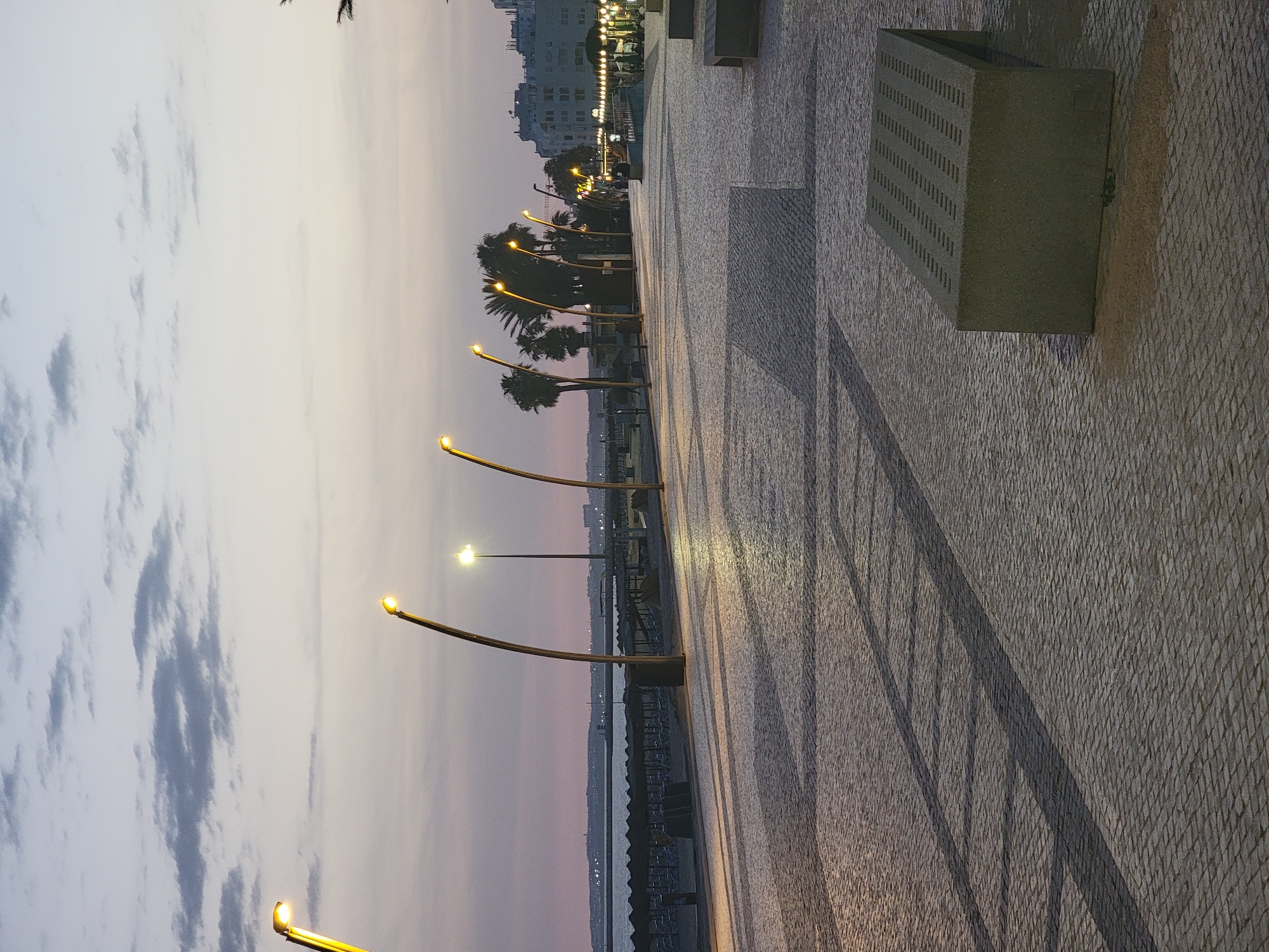 The image depicts a dusk scene at the Quarteira promenade in Portugal. The sky is a soft gradient of pink and grey, with scattered clouds. The promenade itself is paved with cobblestones, with geometric shadows cast by lampposts. A series of modern lampposts with curved, golden poles line the promenade, their lights illuminated. Beyond the promenade, a beach with rows of empty loungers and thatched umbrellas is visible, separated by a low wall. The sea is calm and stretches to the horizon. In the distance, buildings and lights of the city are visible. Palm trees are scattered along the promenade, adding to the tropical atmosphere. A modern, grey concrete bench with a perforated surface is positioned on the right side of the promenade. No people are visible in the immediate foreground. The overall impression is one of a tranquil evening in a coastal resort town.