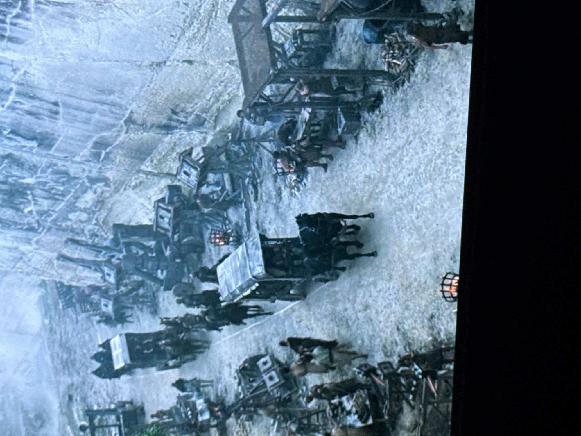 This image, captured from a screen in Sofia, Bulgaria, depicts a fictional or historical scene set in a cold, possibly mountainous or fortified environment.
The scene is dominated by a wide, light-colored path, likely dirt or snow-covered, running from the mid-ground towards the background. To the right of the path, a massive, imposing wall, appearing to be constructed of ice or light-colored rock with vertical striations, extends from the mid-ground into the distance.
Along both sides of the path, numerous makeshift structures resembling market stalls or encampments are present. These structures are primarily dark in color and appear to be constructed from wood or metal. On the right side, at the base of the large wall, several large, dark, complex mechanical structures, possibly cranes or siege engines, are visible.
Numerous individuals, estimated between 20 and 30, are distributed throughout the scene. They are generally dressed in dark, heavy, layered clothing, consistent with a cold climate. Several individuals are riding dark-colored horses, while others are walking on the path or standing at the stalls. Some figures appear to be engaged in activity around the stalls, possibly trading or working.
In the mid-ground, a dark, enclosed horse-drawn carriage, pulled by two dark horses, proceeds along the path towards the viewer's left. Another similar carriage is partially visible further back on the left.
The overall lighting is diffuse and cool-toned, contributing to a somber or cold atmosphere. Colors are muted, primarily consisting of blues, grays, whites, and dark browns. A glowing brazier or fire pit, set within a metal cage, is partially visible at the bottom center of the image, cropped by the screen's edge. The image exhibits characteristics of a photograph taken of a display screen, including potential pixelation and a dark border at the bottom.