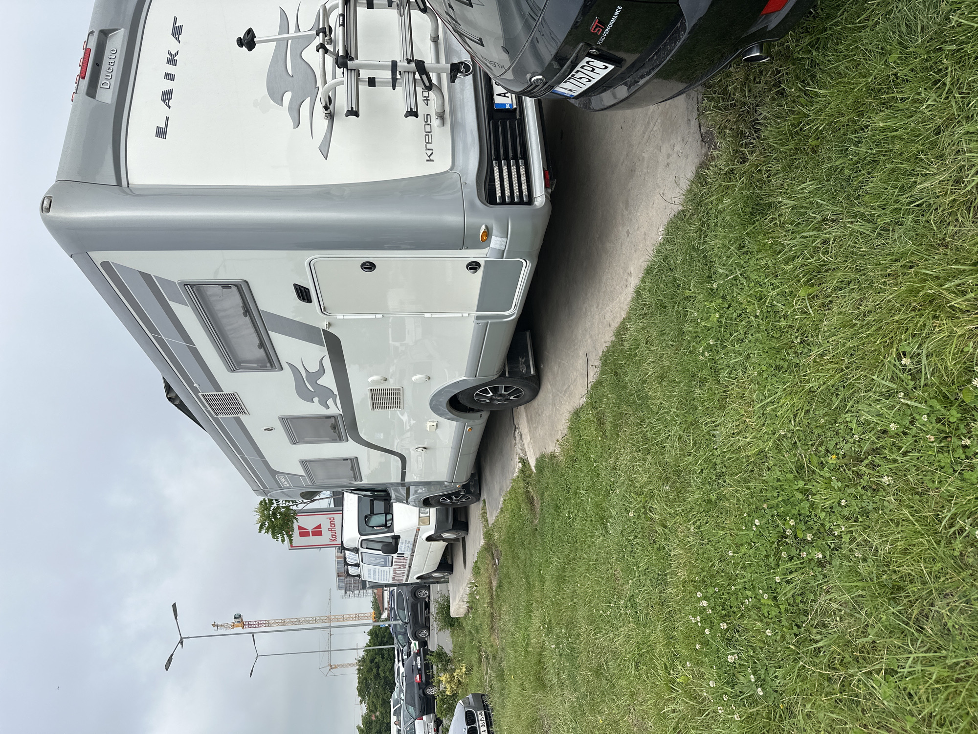 An outdoor, daytime scene shows several vehicles parked alongside a grassy strip, with the sky overcast. The primary subject is a large, grey and white motorhome, a "Laika Kreos 40" built on a "Ducato" chassis, prominently displaying "LAIKA" on its rear. It has a bicycle rack mounted on the back.
Immediately behind the motorhome is the front end of a black "Ford Performance" car, partially visible, bearing the license plate "A 7757 PC" and a "BG" (Bulgaria) sticker. Further down the paved lot, a white van marked with the Cyrillic text "АВТОСТЪКЛА" (Auto Glass) is parked, followed by a line of other cars, one of which shows the license plate "A 0615 HM".
In the background, a "Kaufland" supermarket sign is visible, along with two large construction cranes and some distant buildings, suggesting an urban or suburban setting. The ground to the right of the parked vehicles is covered in green grass with small white clover flowers. The image is taken from a tilted perspective, looking across the vehicles and the grassy area towards the sky and background infrastructure. The overcast conditions suggest a mild or cool day, with no direct sunlight. The presence of Cyrillic script on the van and the Bulgarian license plates indicate the location is likely Bulgaria.
