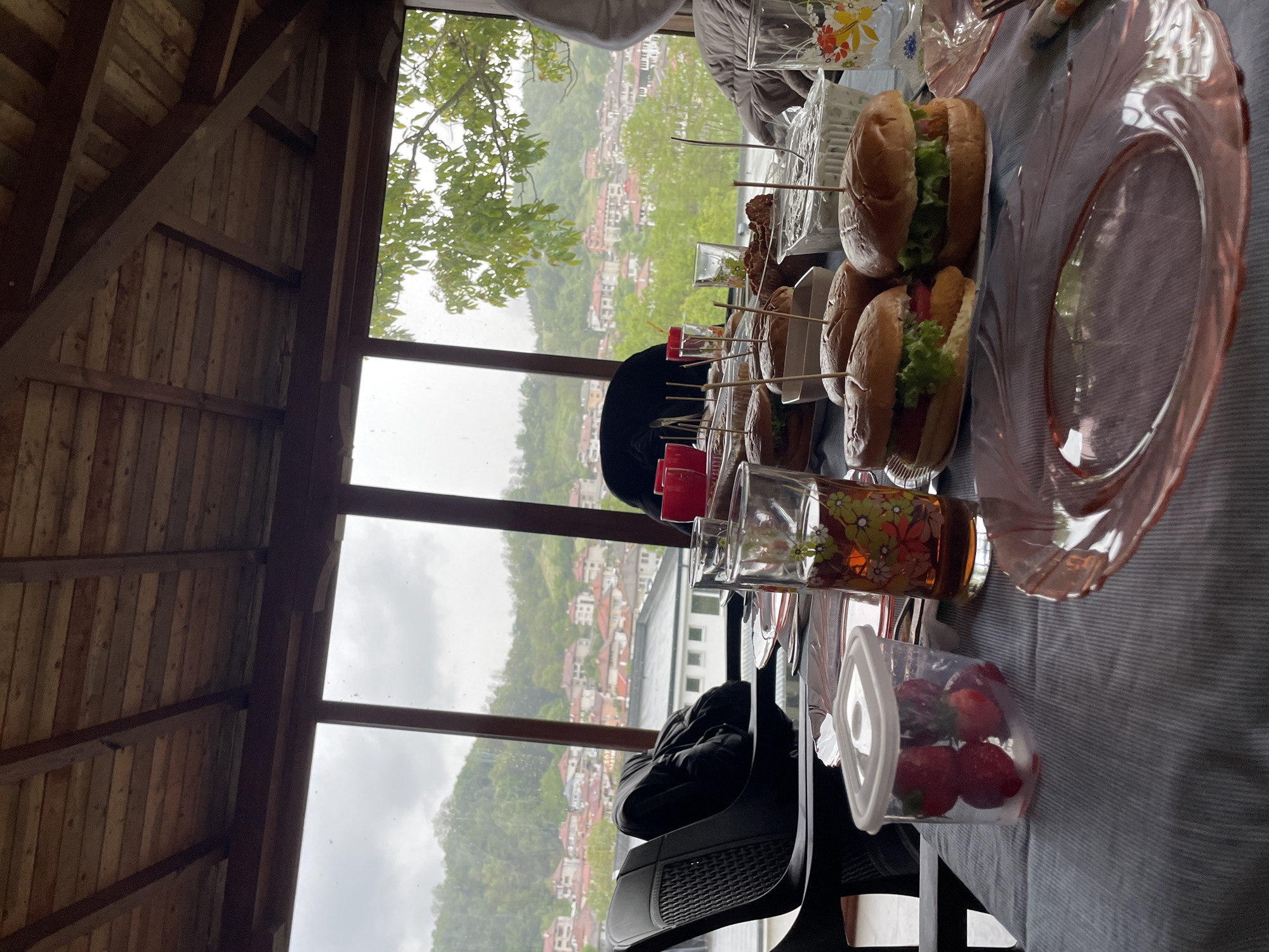 The image shows a table set with food, including sandwiches and strawberries, inside a building with a view of Zlatograd, Bulgaria. The building appears to be a wooden structure with large windows overlooking a townscape with numerous buildings and green hills.