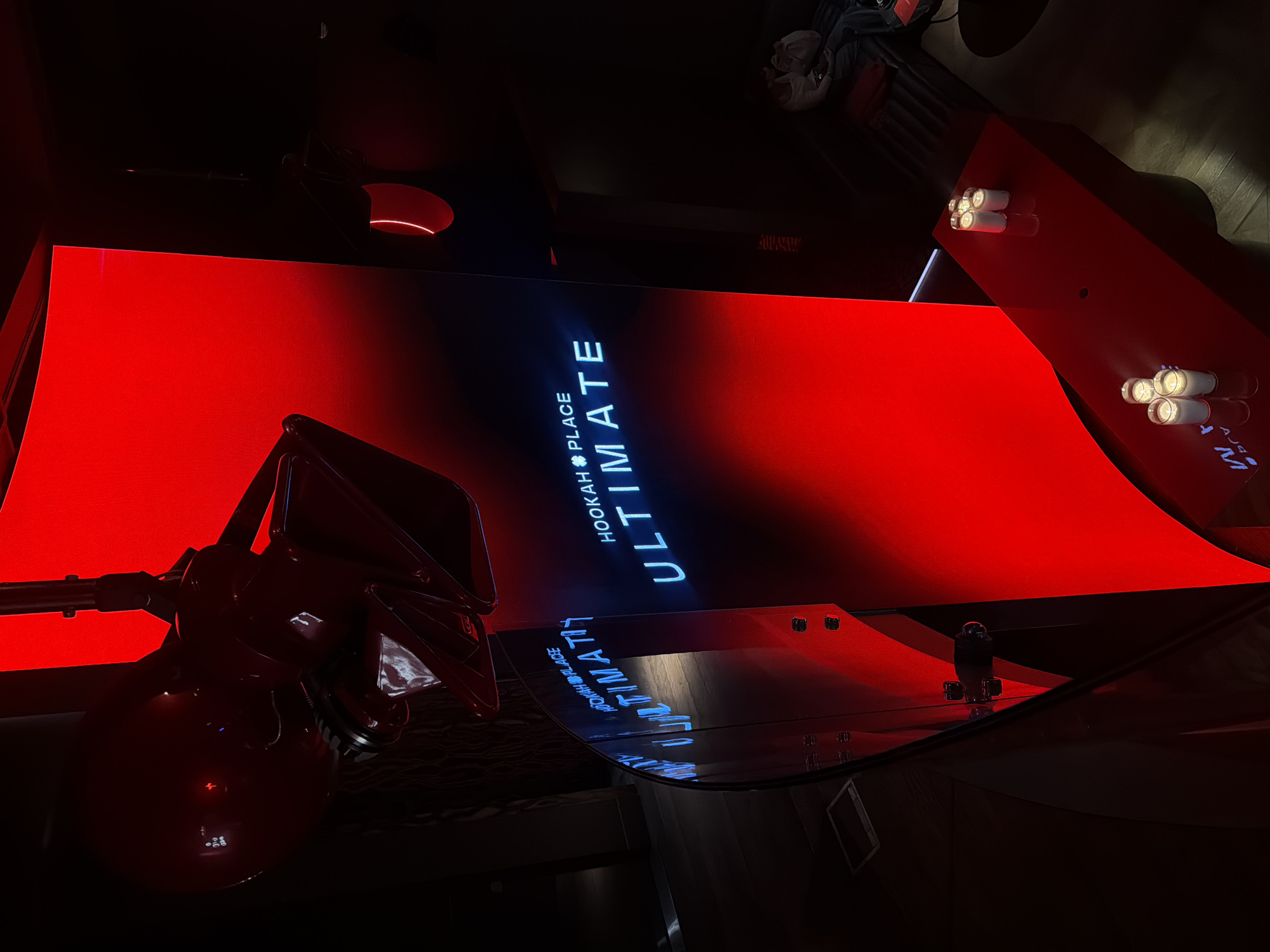 The image presents an interior setting in Moscow, Russia, specifically at a place named "Hookah Place Ultimate." Predominantly red lighting illuminates a modern, stylized hookah lounge. The focal point includes a hookah apparatus in the foreground, positioned near a table or counter surface with the name "Hookah Place Ultimate" displayed in bright blue light. Adjacent to the hookah, a reflective surface mirrors the branding. The backdrop appears to be a large red screen or wall. Further into the background, light fixtures provide additional illumination. The setting suggests a contemporary and possibly upscale establishment.