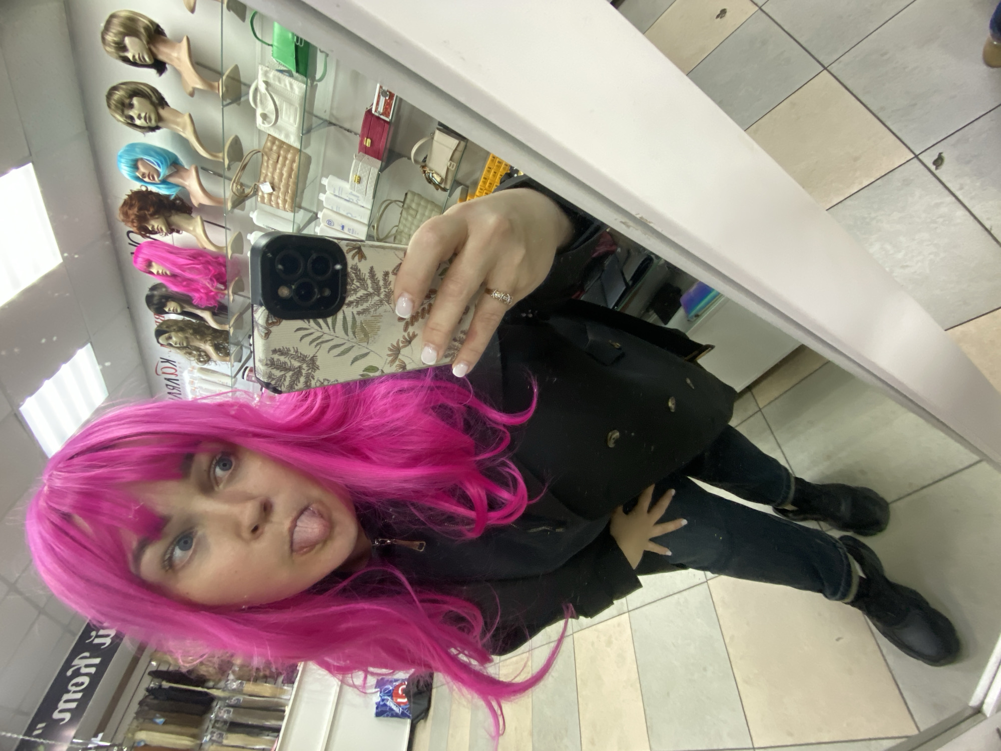 A young woman captures a mirror selfie inside a store, likely a wig or beauty supply shop, in Sochi, Russia. She is trying on a vibrant, long, wavy pink wig with bangs, complementing her blue eyes. She playfully sticks out her tongue towards the camera. She wears a black zipped jacket, dark blue jeans with rolled cuffs, and black flat-soled shoes.
Her left hand holds a silver/gray smartphone with a patterned case featuring green and brown foliage. Her nails have a white-tipped French manicure, and she wears a ring with a double-lobed design on her left ring finger.
The background, visible through the mirror's reflection, displays several wigs on mannequin heads, showcasing various colors including blonde, light blue, reddish-brown, and dark brown. Below the wigs, small bags or purses in cream, green, and red are displayed, along with bottles of what appear to be hair care or cosmetic products on a lower shelf. The floor is tiled with alternating light gray and beige squares. Partially visible, reversed Cyrillic text on a sign in the background reads "ОВЫ", possibly part of a word like "ГОТОВЫ" (Ready) or a store brand name.