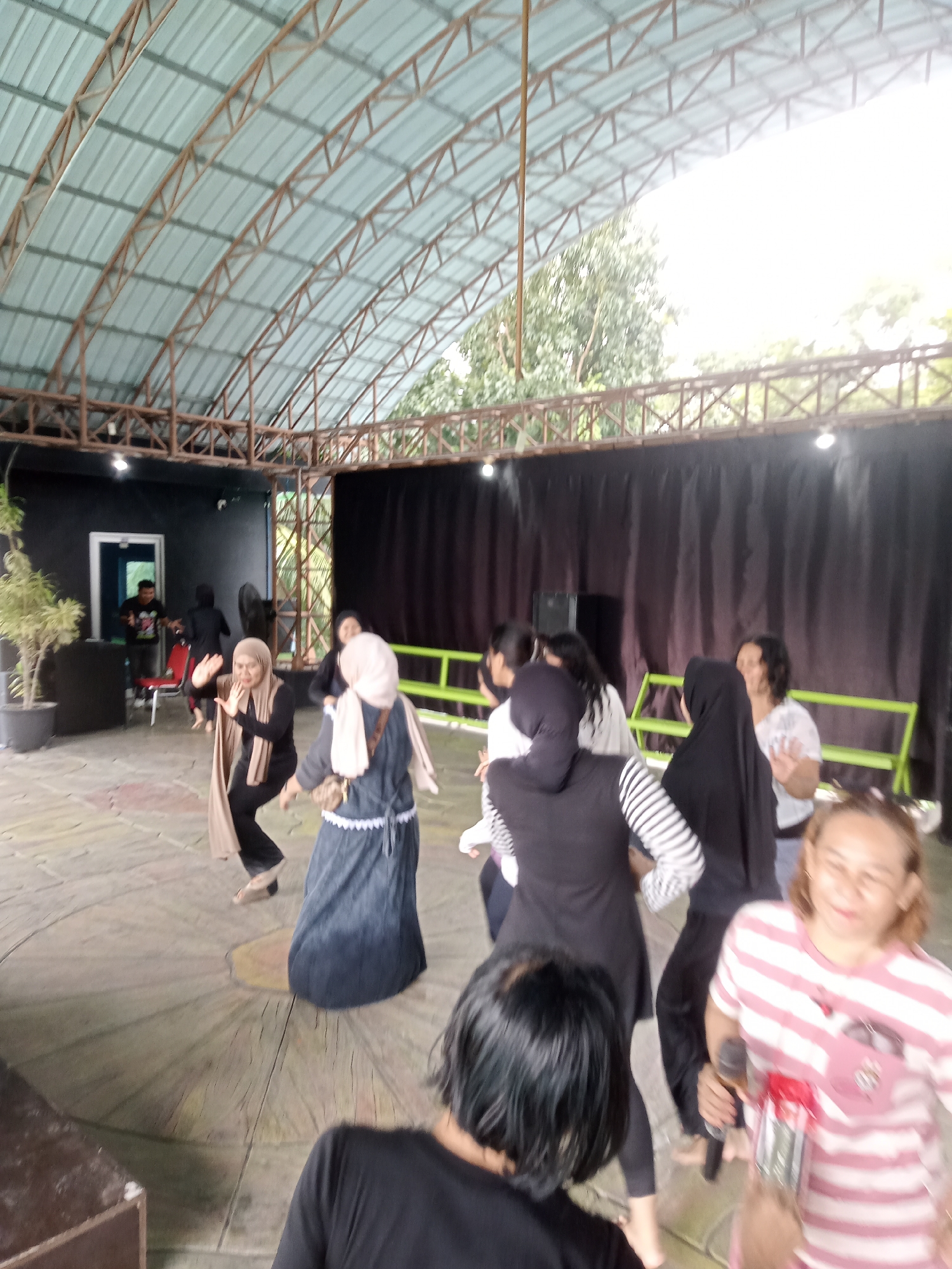 The image depicts an outdoor performance or rehearsal space. A group of people, primarily women, are engaged in an activity that appears to be dancing or practicing a performance. Several individuals are wearing headscarves, and their attire is casual. The setting features a distinctive circular wooden floor with radiating lines, suggesting a stage area. Behind the performers, black curtains create a backdrop, and beyond that, trees and bright daylight are visible, indicating an open-air or semi-open-air venue. A metal roof structure with exposed beams arches over the space. In the foreground, a woman wearing a pink and white striped shirt is holding a microphone and smiling. The image seems to capture a moment of energetic activity and community engagement. Based on the contextual information provided (Baru, Indonesia), this location could be an outdoor cultural center, community space, or a venue for local performances within the city.