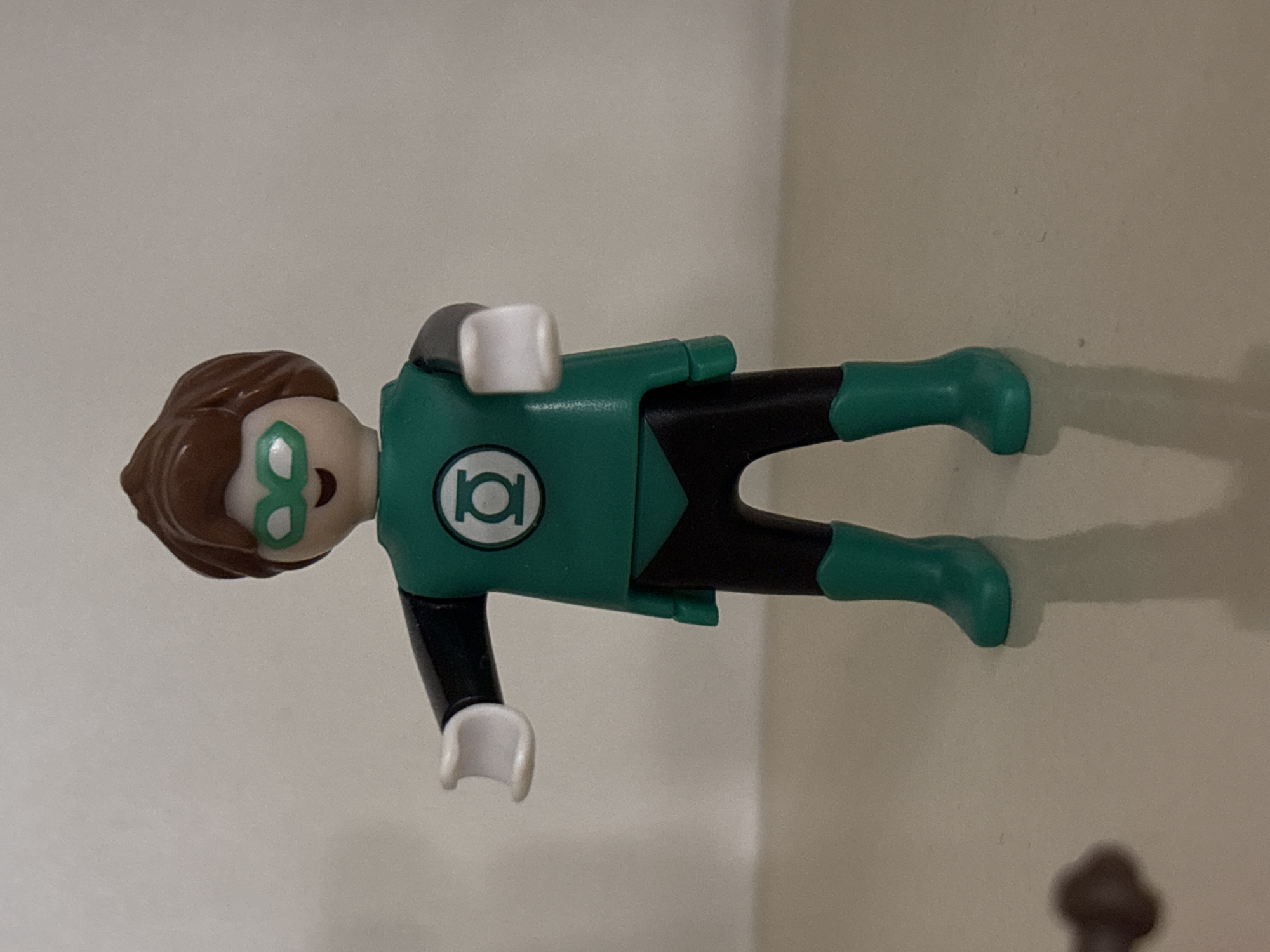 A Green Lantern Playmobil figure is lying on its side on a surface. The figure has brown hair, green glasses, a green torso with the Green Lantern logo, black limbs, and green boots. The background is a plain, light-colored wall. There is no indication of Sofia, Bulgaria, or any specific location.