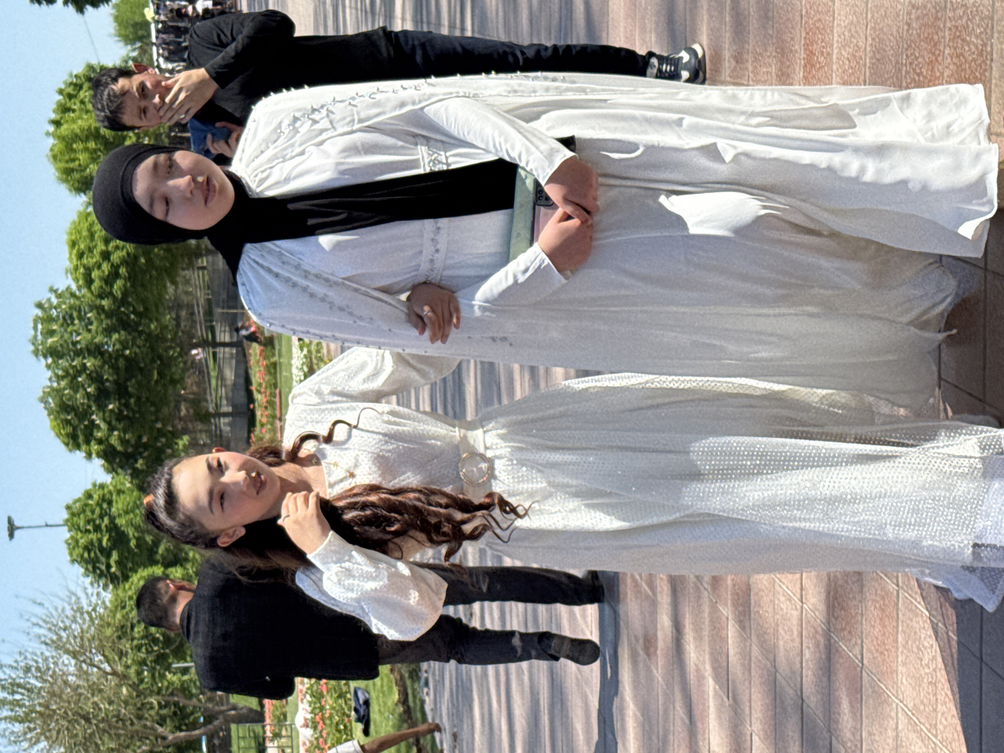 In Samarkand City, Uzbekistan, two women in white dresses stand on a brick pathway in front of a line of trees and bushes with sparse red flowers. The woman on the left has dark brown hair with an orange hair tie and a rhinestone belt. The woman on the right is wearing a black hijab with a long white robe. A man in black is standing to the left of the first woman, and a man in black is behind the second woman, taking a photo. There are people standing behind them, on the pathway.