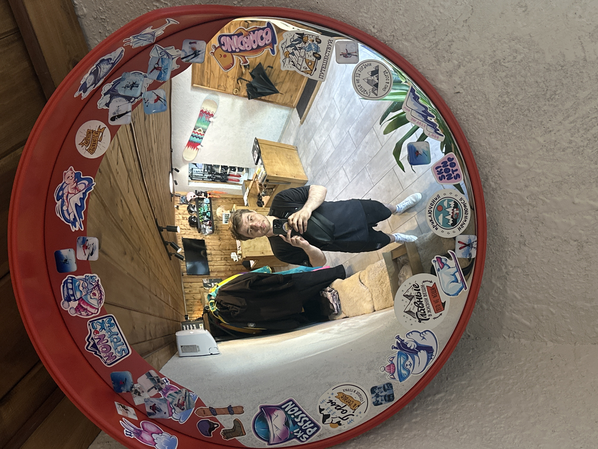 A man is captured taking a selfie in a large, round, convex mirror with a prominent red frame, which is covered in numerous stickers. He is wearing a black t-shirt, dark shorts or pants, and white sneakers, holding a smartphone to capture his reflection. The mirror's curved surface distorts the reflection, making him appear stretched.
The reflection reveals an indoor setting, likely a sports equipment rental shop or a related establishment, in Krasnaya Polyana, Russia. Visible in the background are wooden panel walls, a light-tiled floor, and wooden furniture. A colorful snowboard with a wavy pattern leans against a wall, and an umbrella hangs nearby. Other ski and snowboard gear, including boots and helmets, are also reflected. A green plant and a white appliance (possibly an air purifier) are visible on the floor.
The stickers adorning the mirror and its frame are a notable detail, predominantly featuring themes of winter sports, mountains, and travel. Visible texts include: "SKI PASSION," "Горы в Сердце" (Mountains in Heart), "СЕРДЦЕ В ГОРАХ" (Heart in Mountains), "Главные В ЖИЗНИ ВЕЩИ ЭТО НЕ ВЕЩИ" (The most important things in life are not things), "NON STOP SKI," "приключения начинаются" (adventures begin), "МЕЧТЫ СБЫВАЮТСЯ" (Dreams come true), "ПУТЕШЕСТВУЙ" (Travel), "Счастье не за горами Оно в горах" (Happiness is not far off. It is in the mountains.), and "BOARDING."