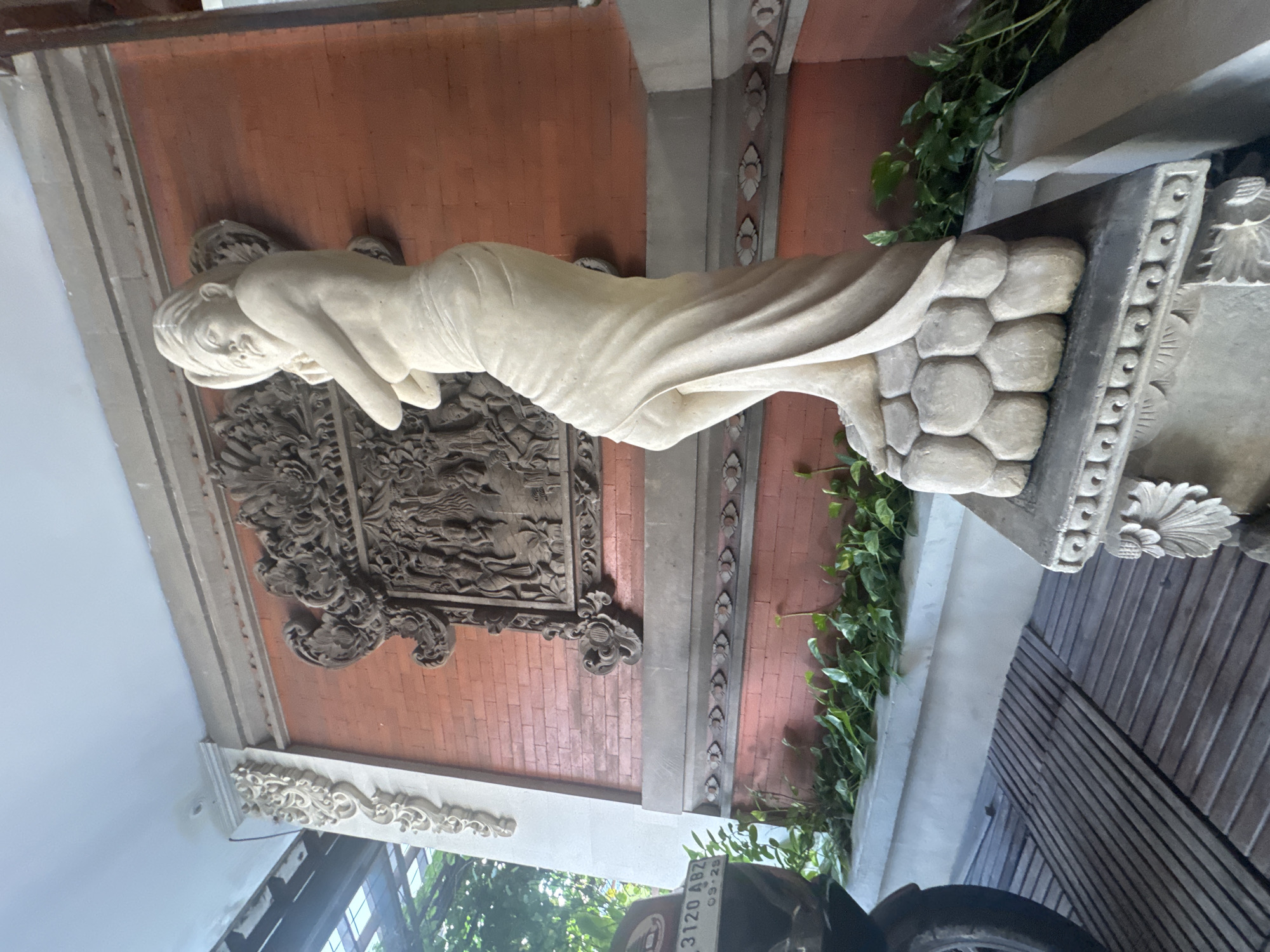 A street-level, medium shot features a reclining statue of a woman placed against a decorated brick wall, likely in Kuta, Indonesia. The woman is sculpted in a pale stone, draped in fabric, and reclines atop a pillar-like structure with a honeycomb pattern and decorative carvings. Behind the statue, the brick wall is adorned with ornate wood carvings and trim. To the side, lush green plants cascade down the wall. A partial view of a vehicle, including a tire and part of a license plate, is visible in the foreground. The overall scene is bathed in natural light.