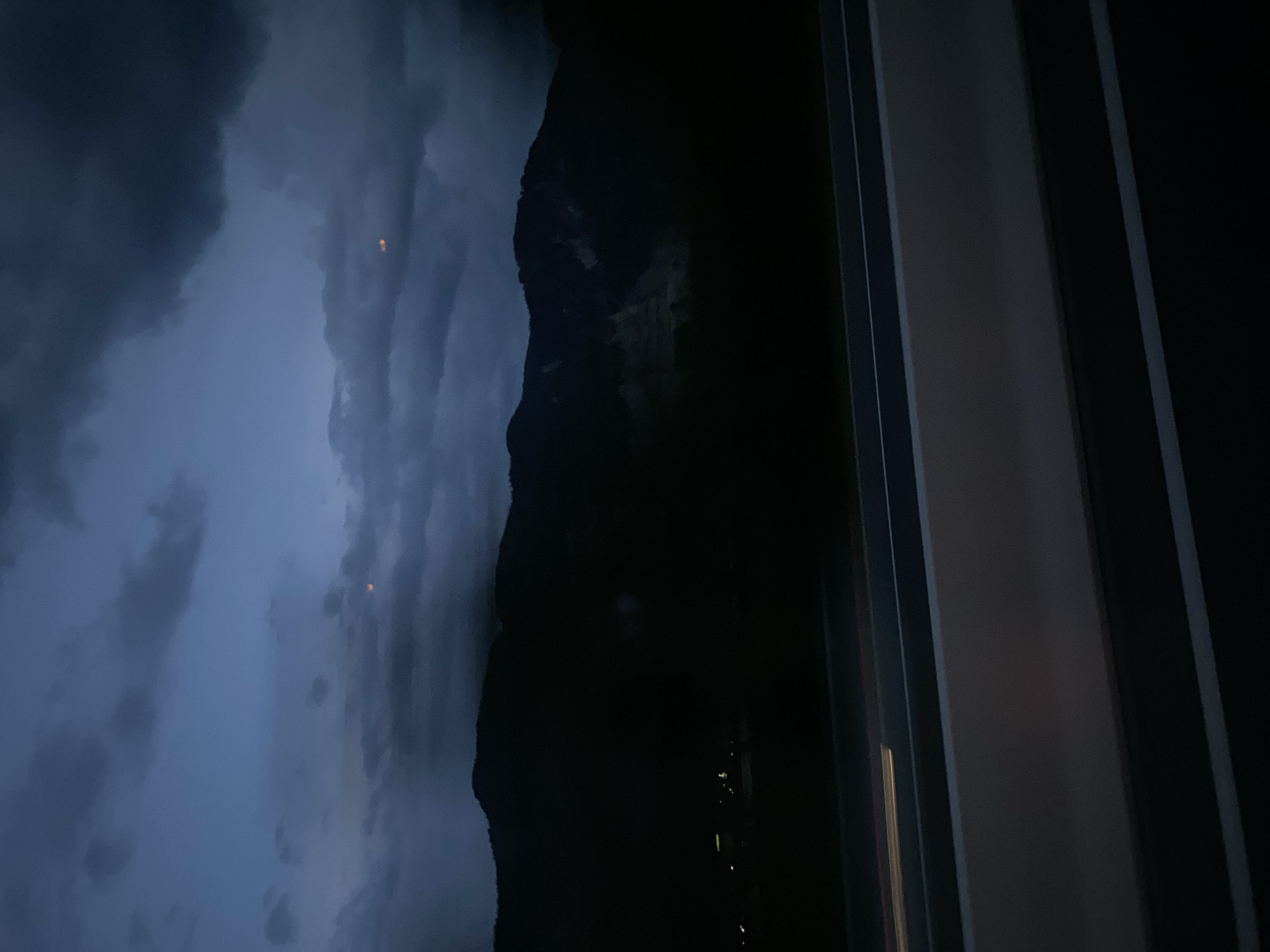 This image captures a dark, cloudy evening or night sky over a mountainous landscape, with a portion of a highway in the foreground. The sky is a deep blue-gray, with heavy, dark clouds obscuring most of the light. Two small, indistinct orange lights are visible in the sky, possibly aircraft or distant lights.
The mountains are silhouetted against the dark sky, appearing as dark masses with some lighter areas that might indicate exposed rock or a quarry. The lower slopes of the mountains descend into deep shadow, making it difficult to discern details. In the far distance, a faint cluster of yellow lights suggests a settlement or town.
The foreground consists of a section of a highway. The highway appears to be multi-lane, with a gray barrier or median strip running horizontally across the lower portion of the image. There are white lines marking the lanes. A streak of light, possibly from headlights of a passing vehicle, is visible on the left side of the highway, indicating motion.
The overall impression is one of a twilight or night scene, likely from a moving vehicle on a highway, looking out towards a mountainous region under a heavy cloud cover. The location in Kundl, Austria, would place this scene in a mountainous area of the Austrian Alps. The specific details of Kundl itself, such as specific buildings or landmarks, are not discernible in this image due to the low light conditions and the focus on the landscape and sky.