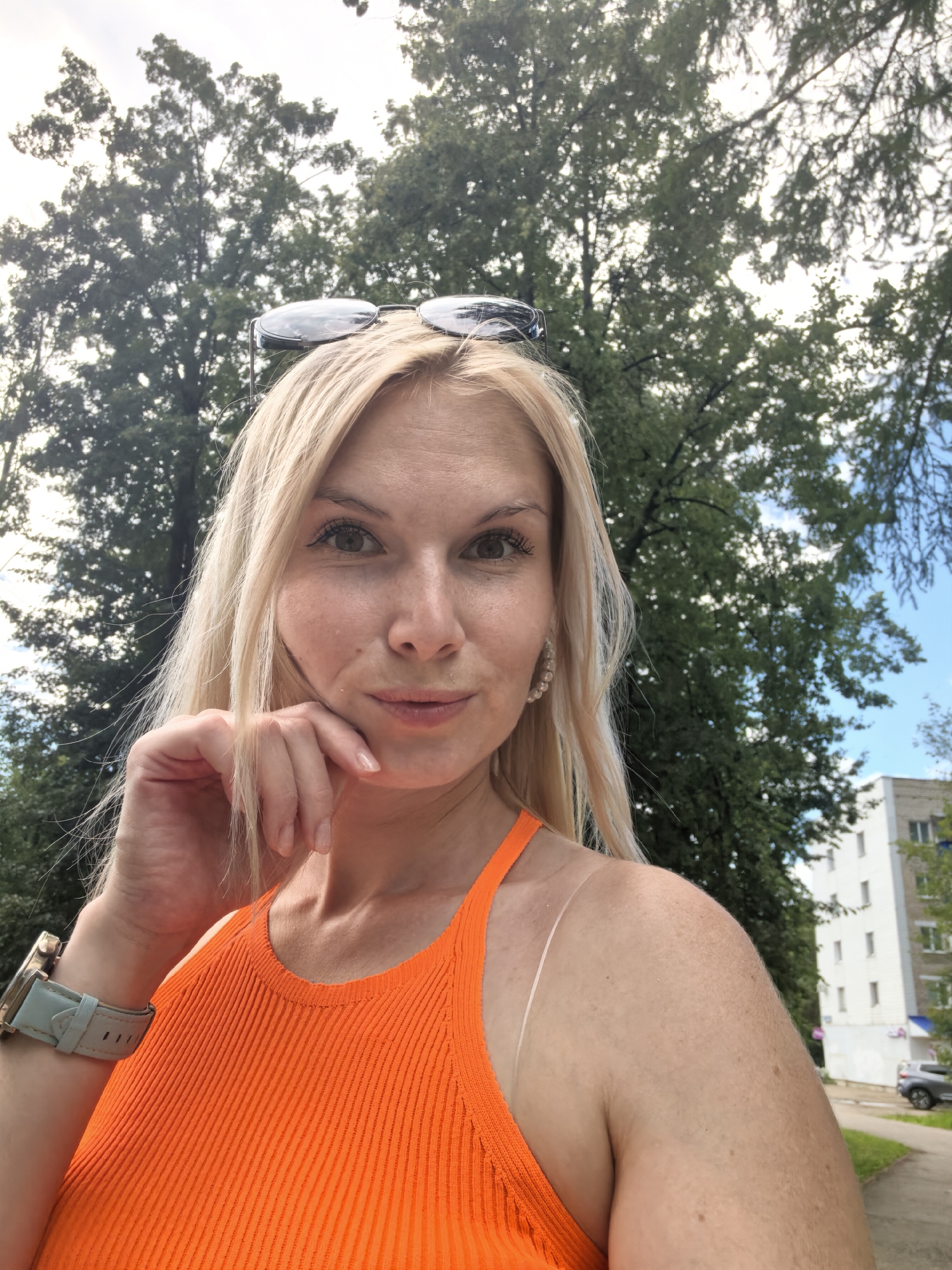 A blonde woman with sunglasses perched on her head poses for a selfie outdoors. She is wearing an orange sleeveless top and a watch on her left wrist. Her right hand is resting on her chin. The background shows trees and a building. The scene is set in Sterlitamak, Russia, as indicated by the context.
