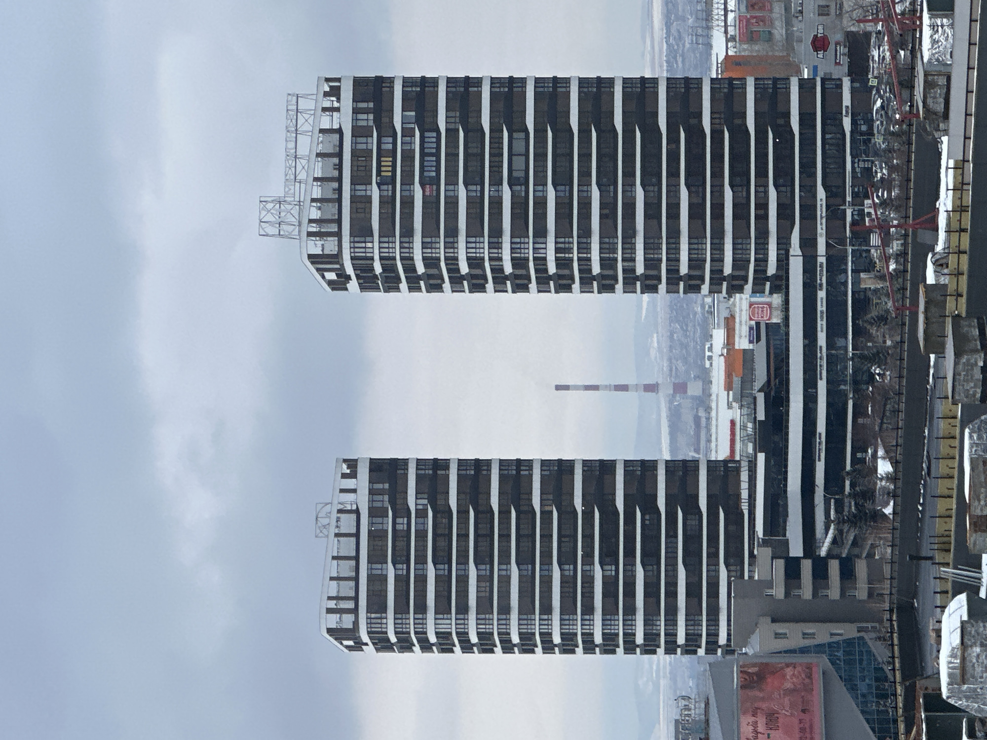 The image captures a panoramic view of an urban landscape in Krasnoyarsk, Russia, under an overcast sky. Two prominent, modern high-rise buildings, identical in design, stand in the mid-ground. Each building features a dark facade with regularly spaced vertical elements that appear to be light-colored, creating a striped pattern. The buildings have multiple stories of rectangular windows and a distinctive structural framework on their roofs.
In the background, a sprawling cityscape is visible, with a mix of residential and commercial structures, and a tall, red and white striped industrial chimney rising above the distant horizon. The general impression is one of a dense urban environment, possibly extending to a river or plain in the far distance, though the details are hazy due to the weather.
The foreground on the right reveals a construction site with red crane structures and exposed building materials, suggesting ongoing development. Between the main buildings and the construction site, several vehicles are parked in what appears to be a lot or street.
The weather is cloudy, lending a muted, cool light to the scene. The overall color palette is dominated by greys, whites, and dark building tones, indicating a chilly day, possibly late autumn or winter, though no snow is clearly visible on the ground.
Visible text includes a circular Burger King logo on a building in the mid-ground, and the text "AUTO.RU" on another building, likely referring to a Russian online car marketplace. Another sign in the lower left foreground (in original orientation) advertises "Брази́льская му́зыка" (Brazilian music) and "КИНО" (CINEMA/MOVIE), indicating a cultural event or film. No people are explicitly visible in the scene.