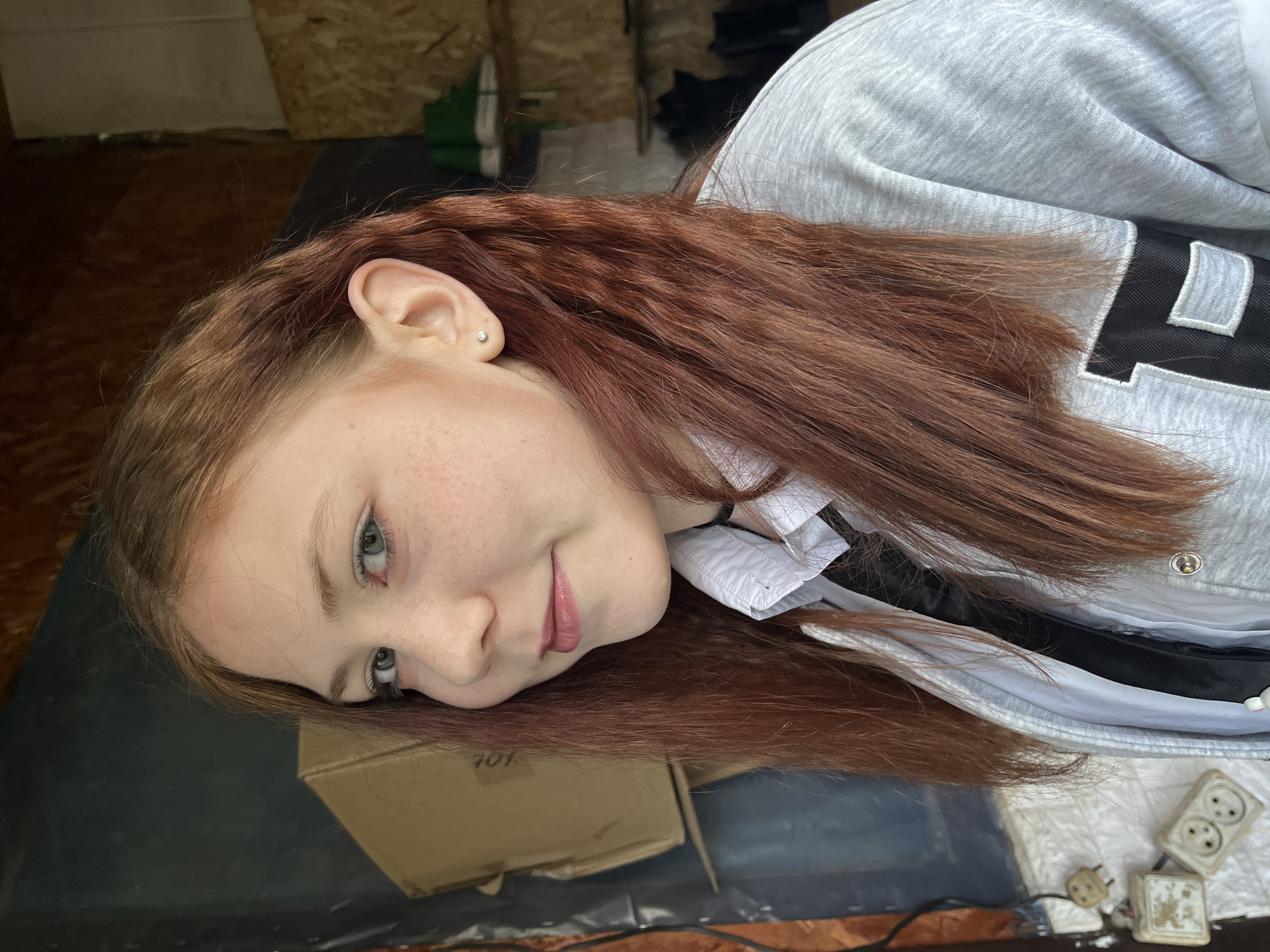 This is a close-up portrait of a young person, likely female, with long, wavy reddish-brown hair, seen from the chest up. Her head is tilted, resting on a dark surface, and she gazes towards the camera with a gentle, subtle smile. She has light blue-green eyes and light freckles scattered across her nose and cheeks. A small, sparkling stud earring is visible in her earlobe. Her lips have a soft pink tint.
She is wearing a light grey hooded sweatshirt featuring a distinct black and white letter "L" design with a white outline on the sleeve. A white collared garment is visible underneath her sweatshirt.
The setting appears to be an indoor space, possibly a rustic or utilitarian room, with wooden planks visible on the wall or ceiling in the background. The surface she rests her head on is dark, potentially a covered floor or table. To the lower right of the image, several old-fashioned electrical outlets and plugs are visible, suggesting an older installation. Part of a cardboard box is also visible in the lower left, displaying the faint numbers "701" on its side.
The lighting is natural and soft, characteristic of daytime. The overall emotion conveyed by the subject is calm and relaxed. The scene is located in Vesyoloye, Russia.