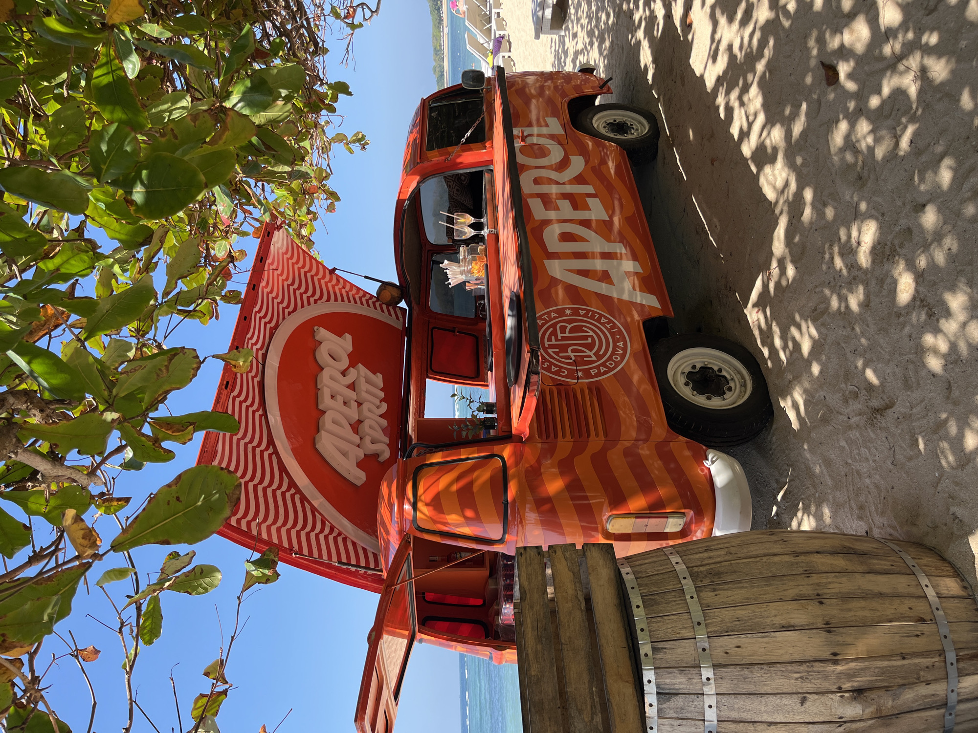 An orange Aperol Spritz van is parked on a sandy beach in Kuta Selatan, Indonesia. The van is adorned with wavy orange and white stripes and features an open serving window. Two glasses with straws sit on the counter of the van. In the foreground, a wooden barrel stands partially visible. In the background, the ocean and lounge chairs can be seen. The scene is shaded by the leaves of a tree.