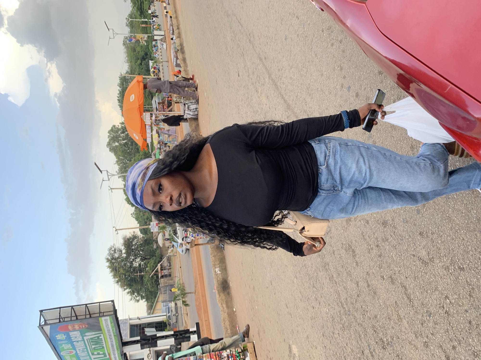 A young Black woman with long, curly black hair and a blue and white headscarf walks on a paved road in Jos, Nigeria. She is wearing a black long-sleeved shirt and blue jeans, carrying a beige shoulder bag and a white plastic bag in her right hand, and a smartphone in her left. The background shows a busy street with vendors, vehicles, and a billboard advertising a political figure. The sky is cloudy.