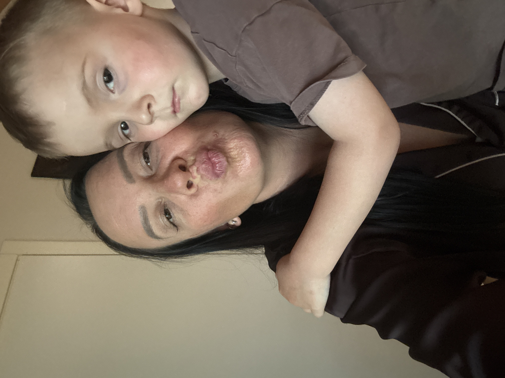 A selfie depicts a woman and a young child. The child is on top of the woman, possibly hugging her. Both are wearing brown clothing. The woman is pouting or making a kissing face. The background is a plain wall. The location is unknown, but the user requested that it be specified as the city of Chaikovsky, Russia.